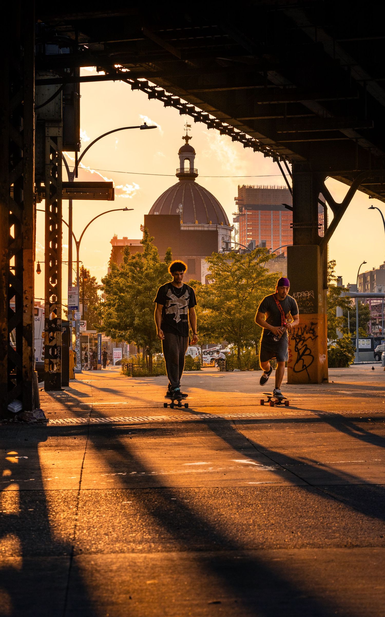 Two skateboarders riding beneath an elevated subway in Williamsburg, Brooklyn during golden hour.