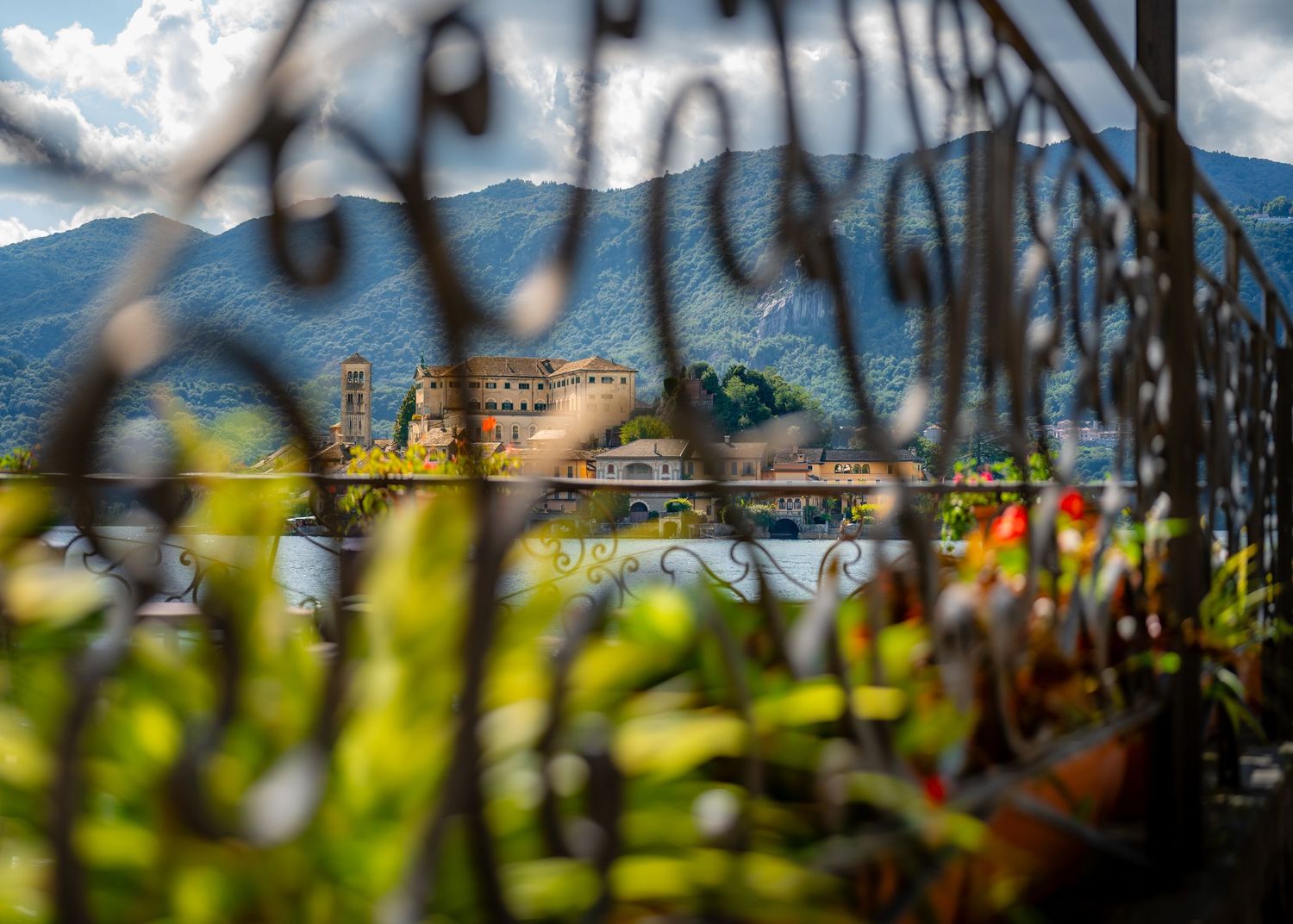 Green plants framing a view of Lake Orta, Italy, and its island, with mountains rising in the distance.