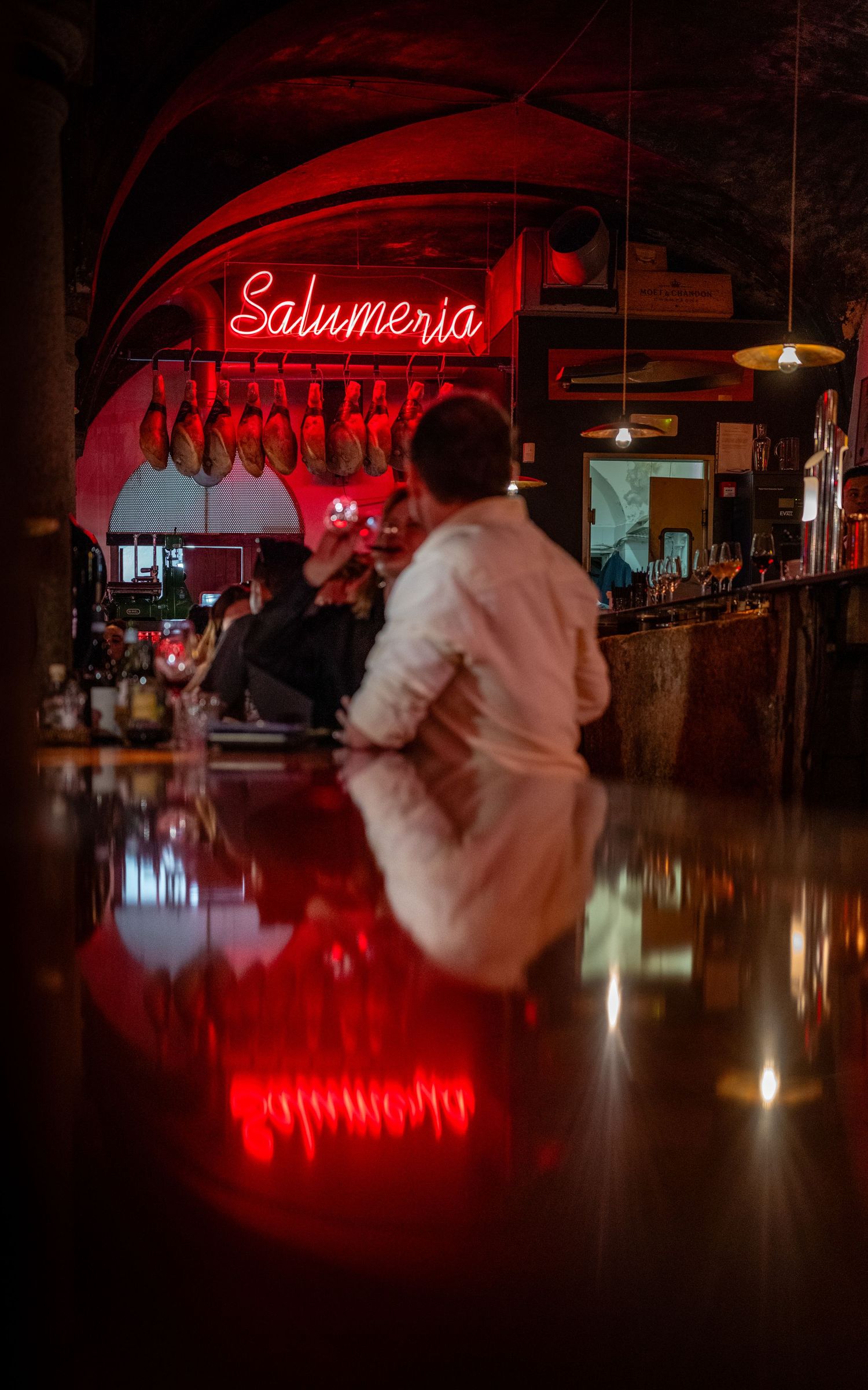 A couple standing at an aperitivo bar in Milan, reflected in a glossy table beneath red neon lights.