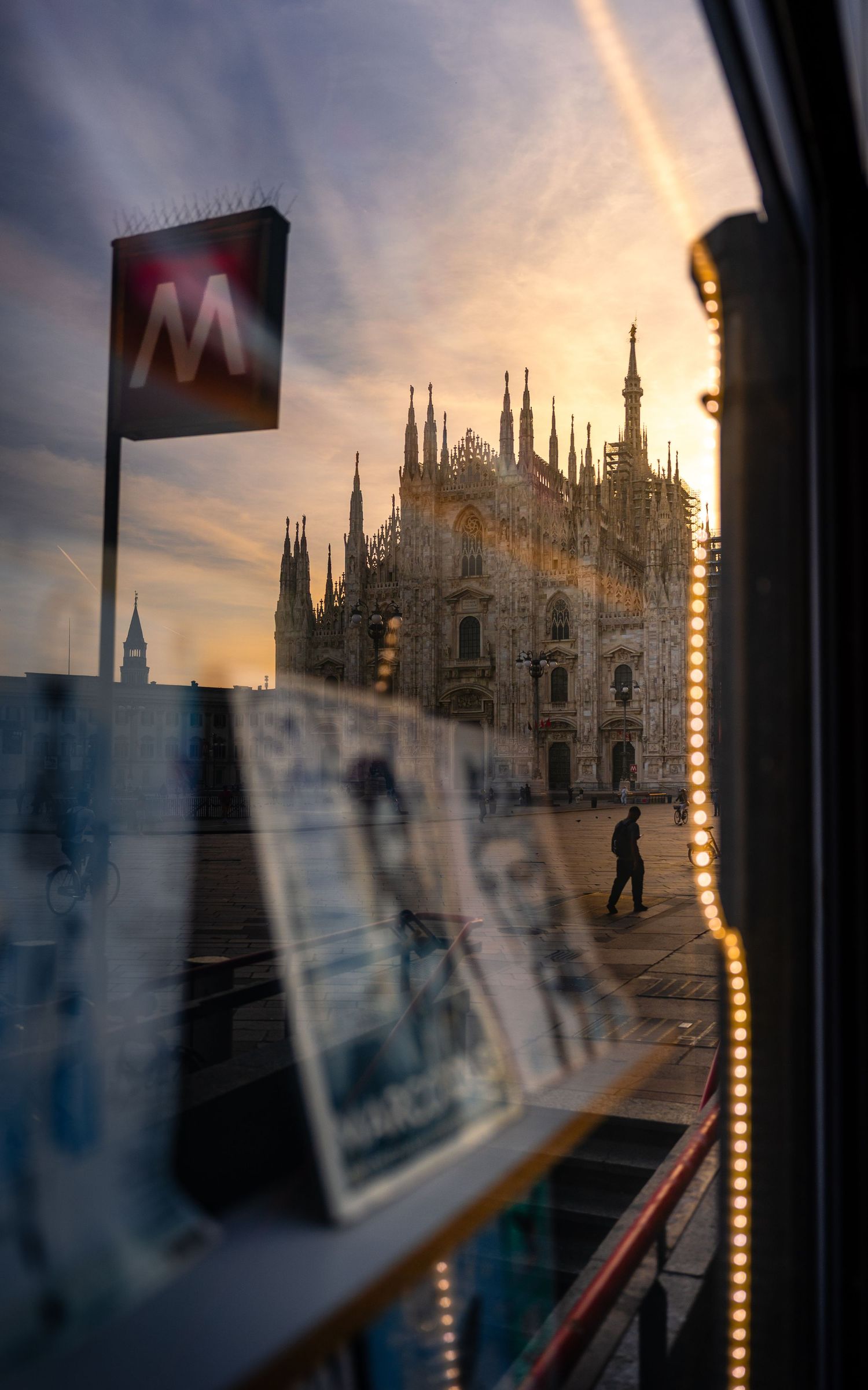 Reflection of Milan’s Duomo in a newsstand window, with the metro sign visible above.