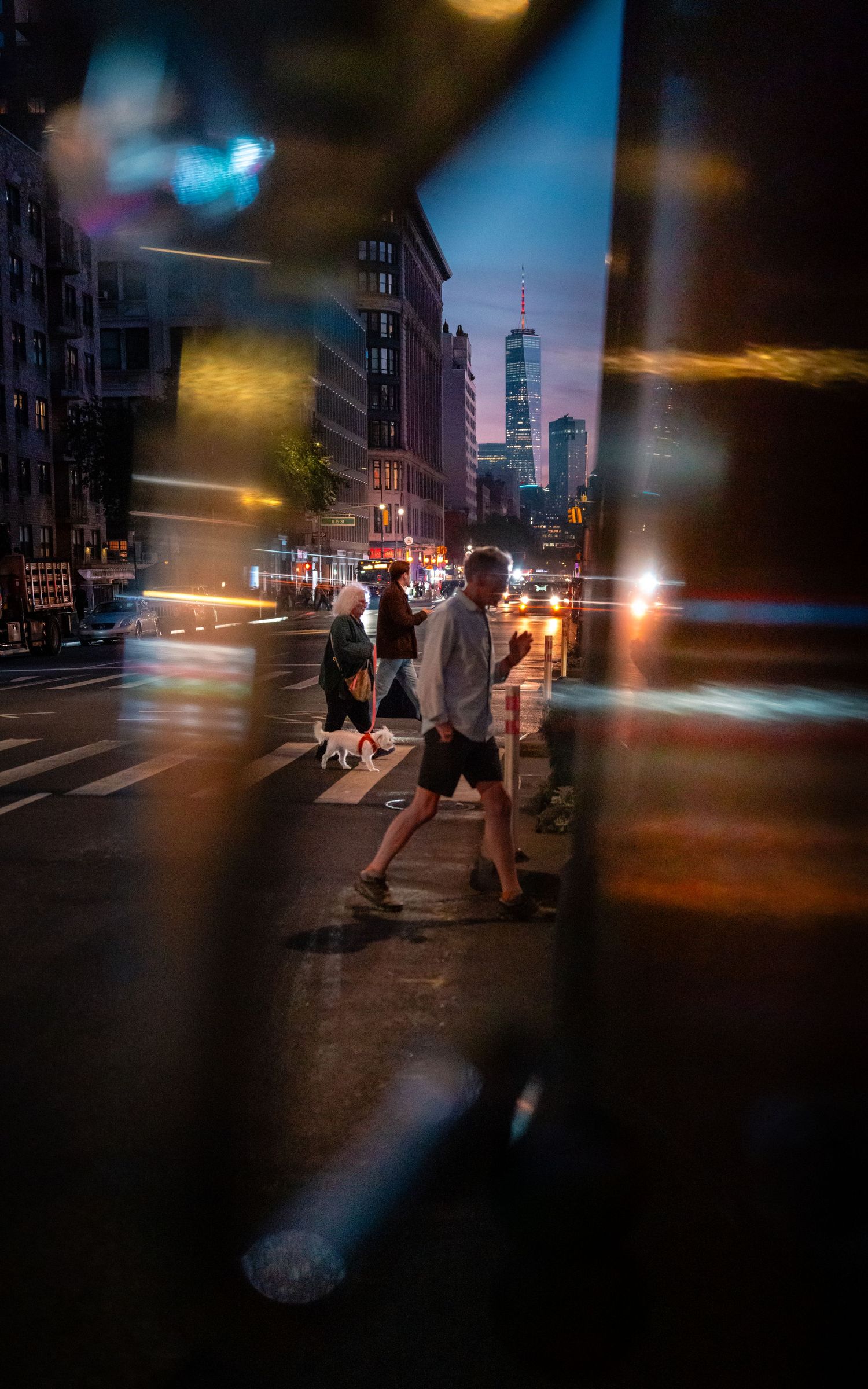 Pedestrians crossing a New York City street at night, with blurred headlights and streetlights streaking through the scene.