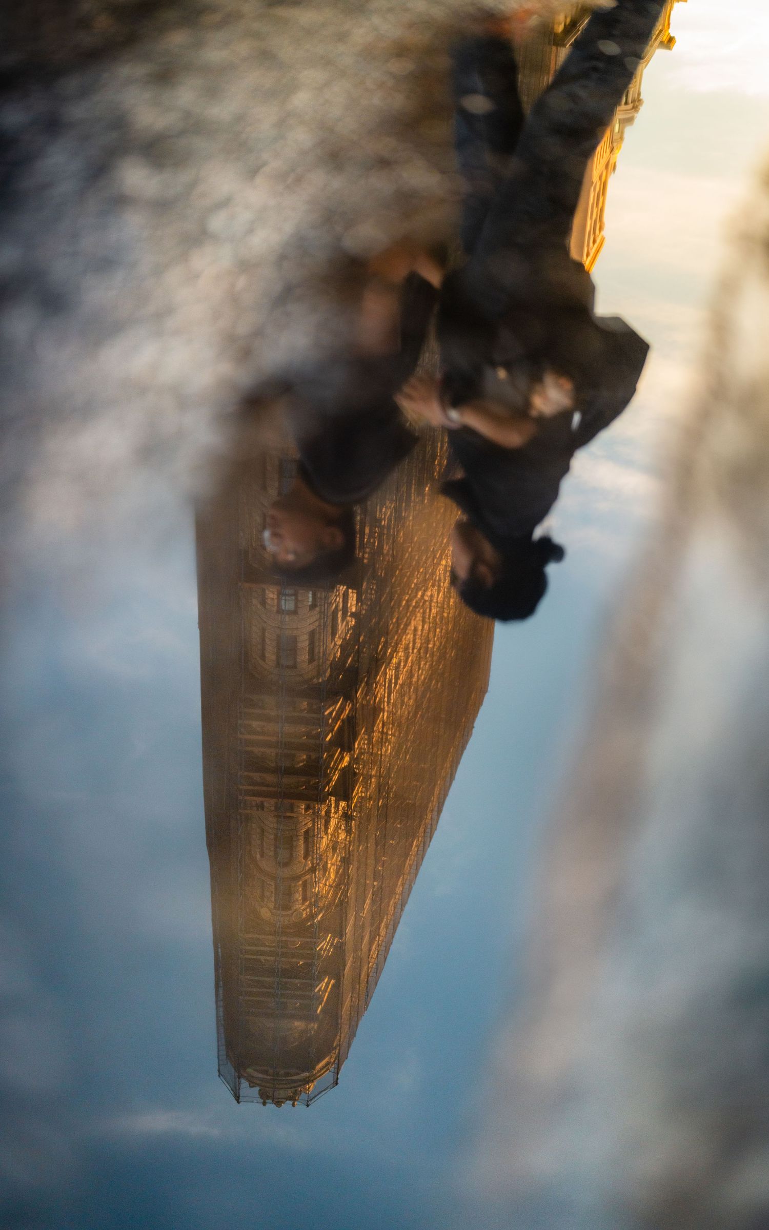 Upside-down reflection of a couple walking past the Flatiron Building mirrored in a puddle.
