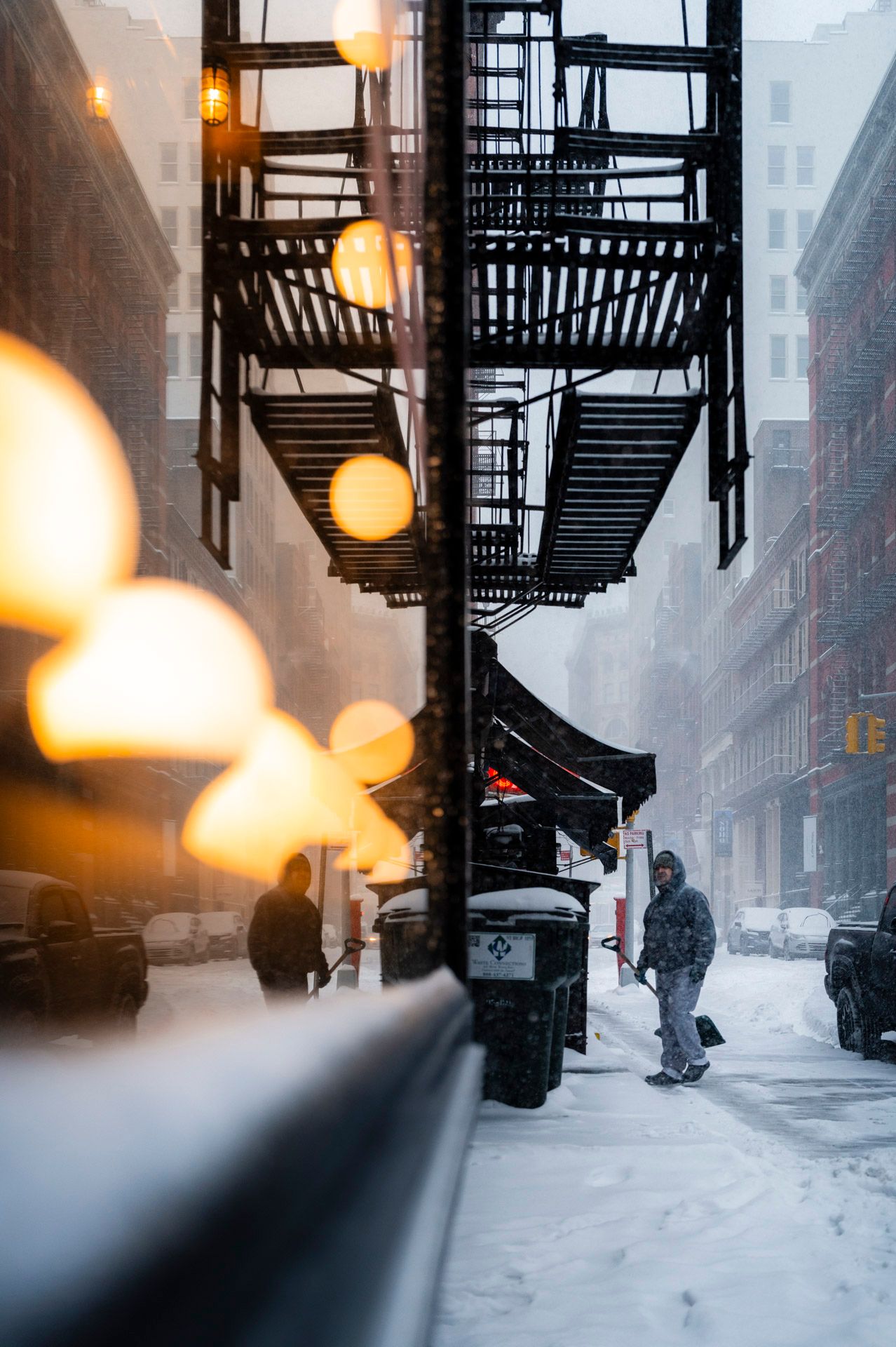 Worker shoveling snow on a SoHo street framed by fire escapes and warm window reflections