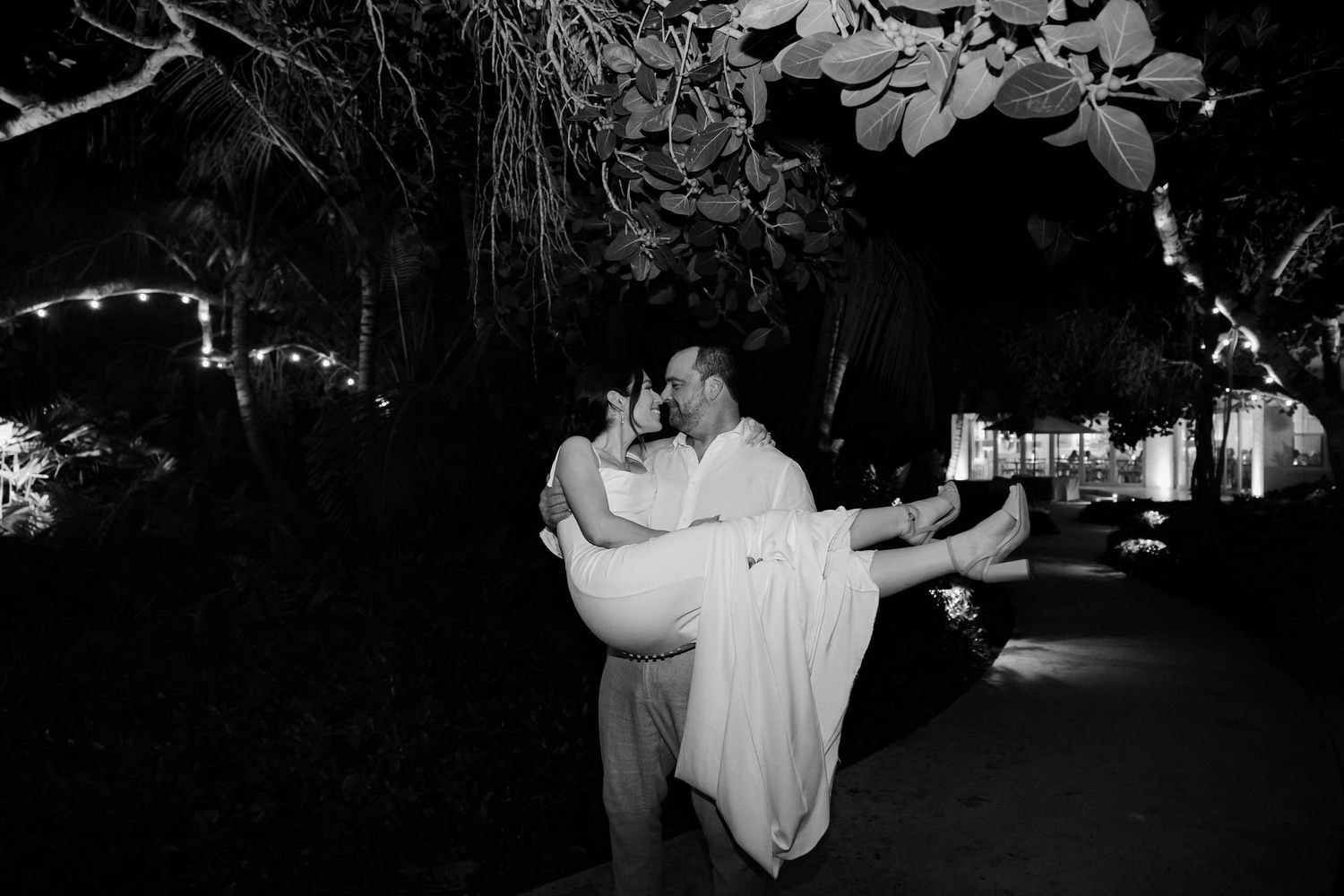 Black and white photo of groom carrying bride in wedding attire outdoors at night with string lights in background.