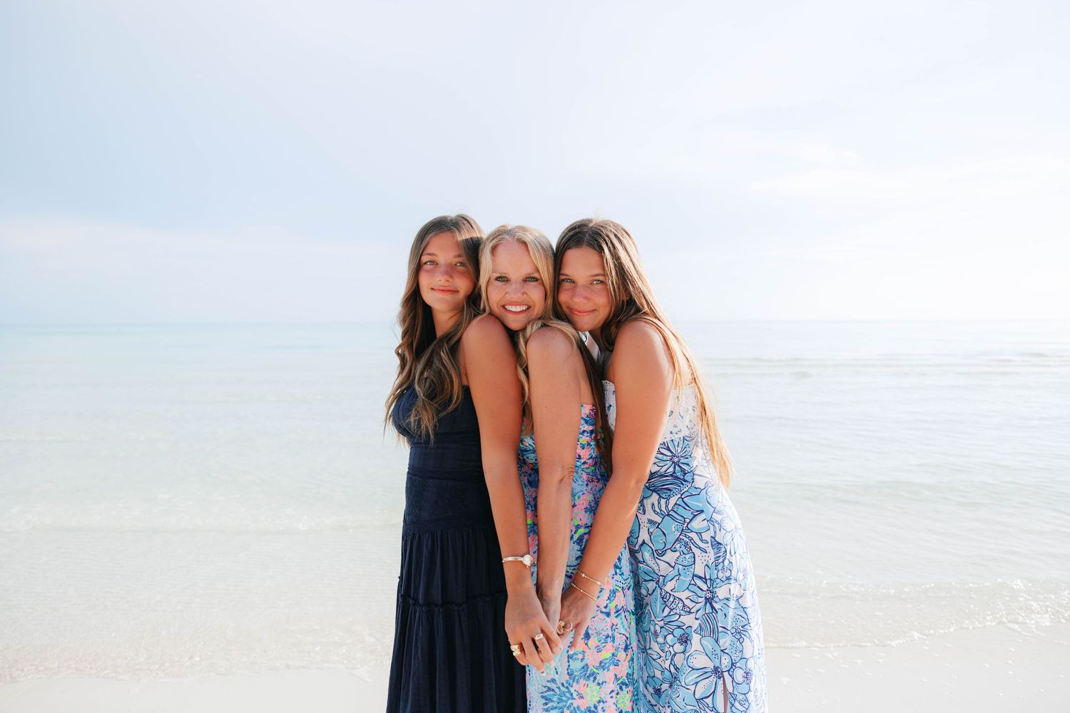 Three girls in summer dresses smiling together on a sunny beach with calm ocean waves in the background.