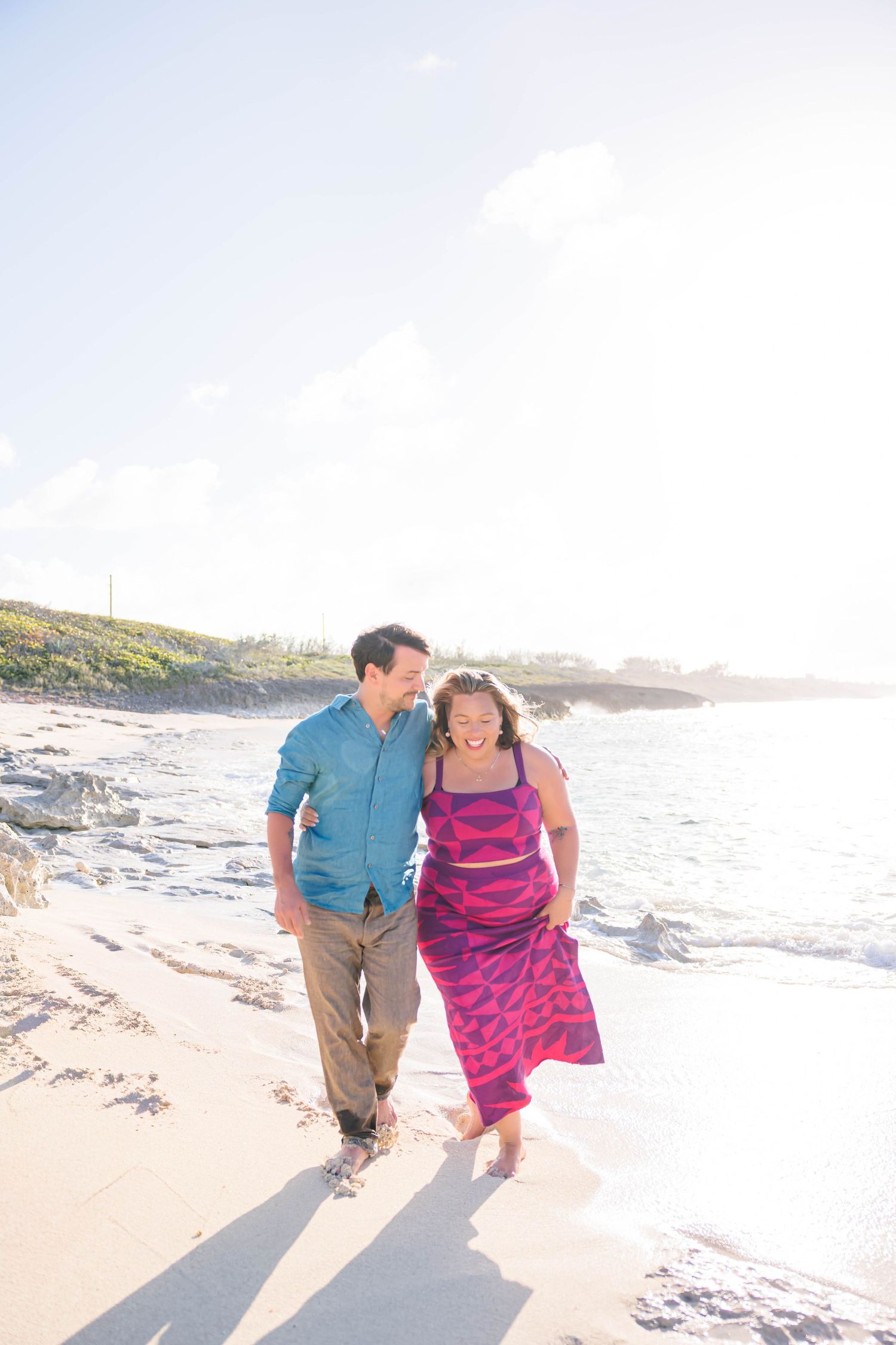 Couple walking barefoot on a sunny beach, man in teal shirt and woman in pink dress, laughing together by the ocean.
