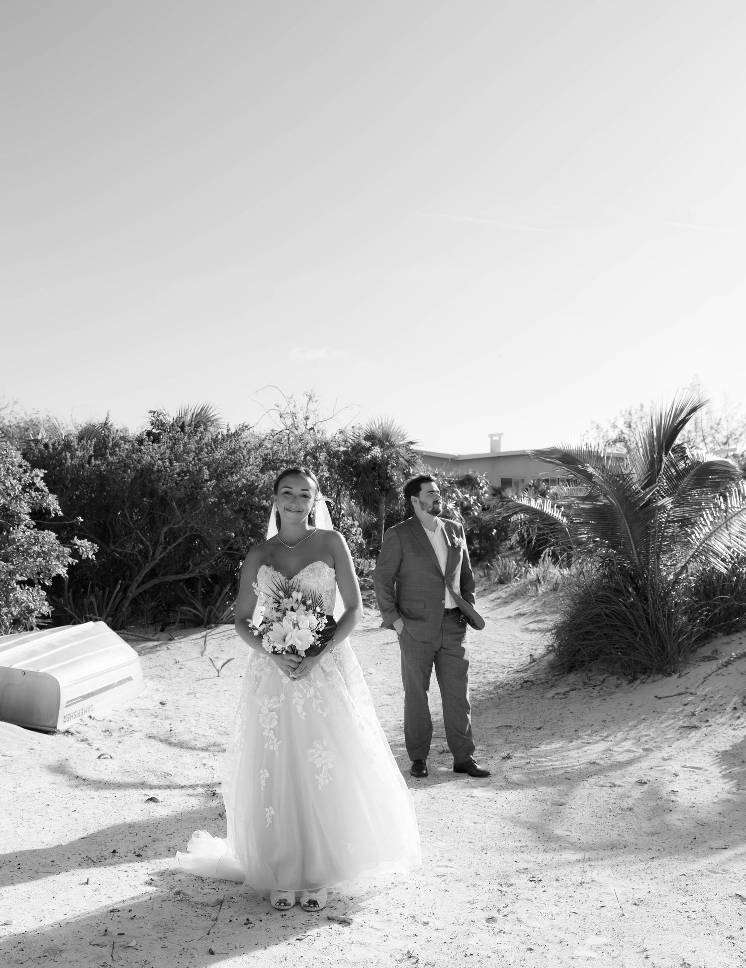 Black and white beach wedding photo of bride in ballgown holding bouquet with groom on sandy path.
