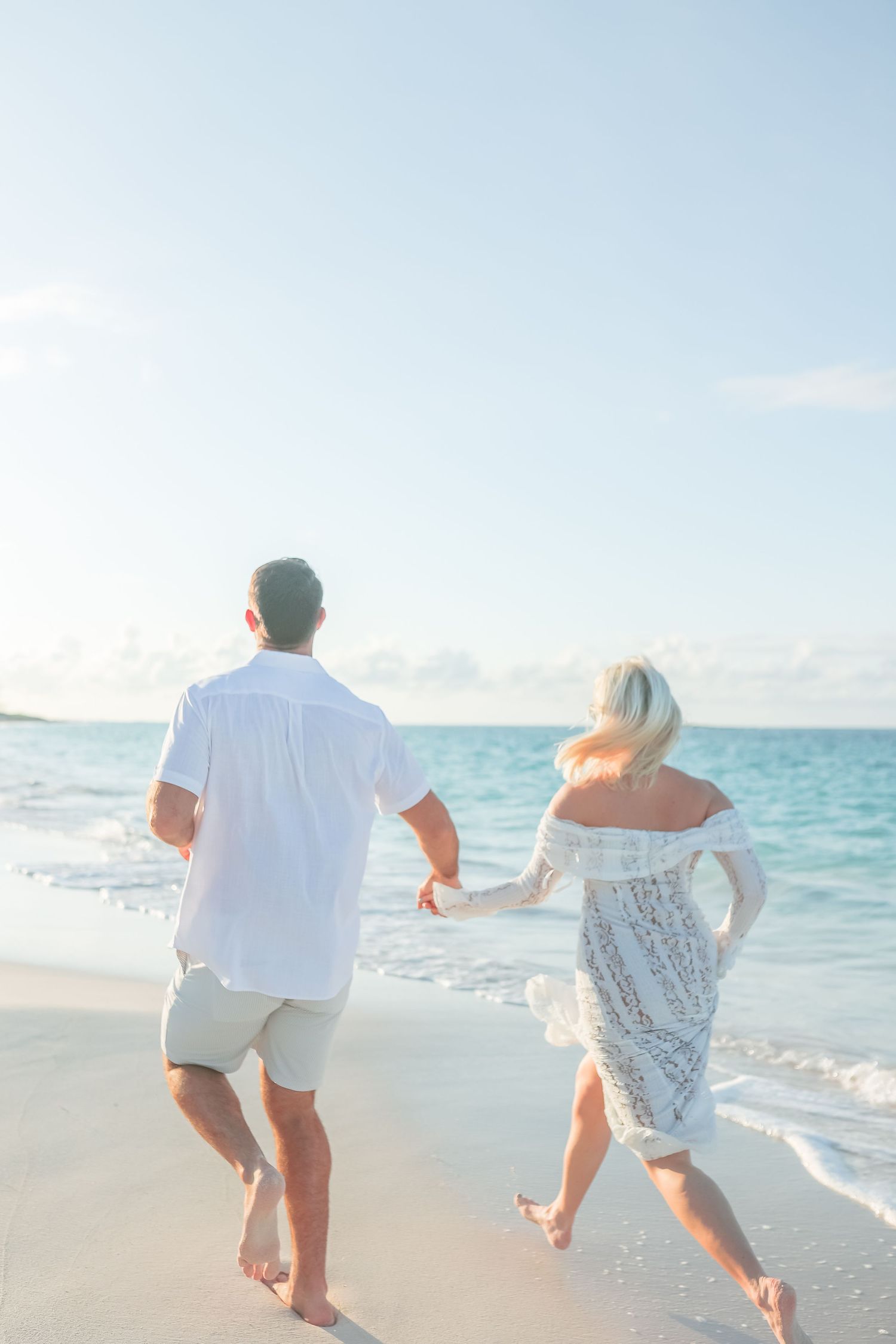 Couple in white outfits holding hands running barefoot on a tropical beach with turquoise water.