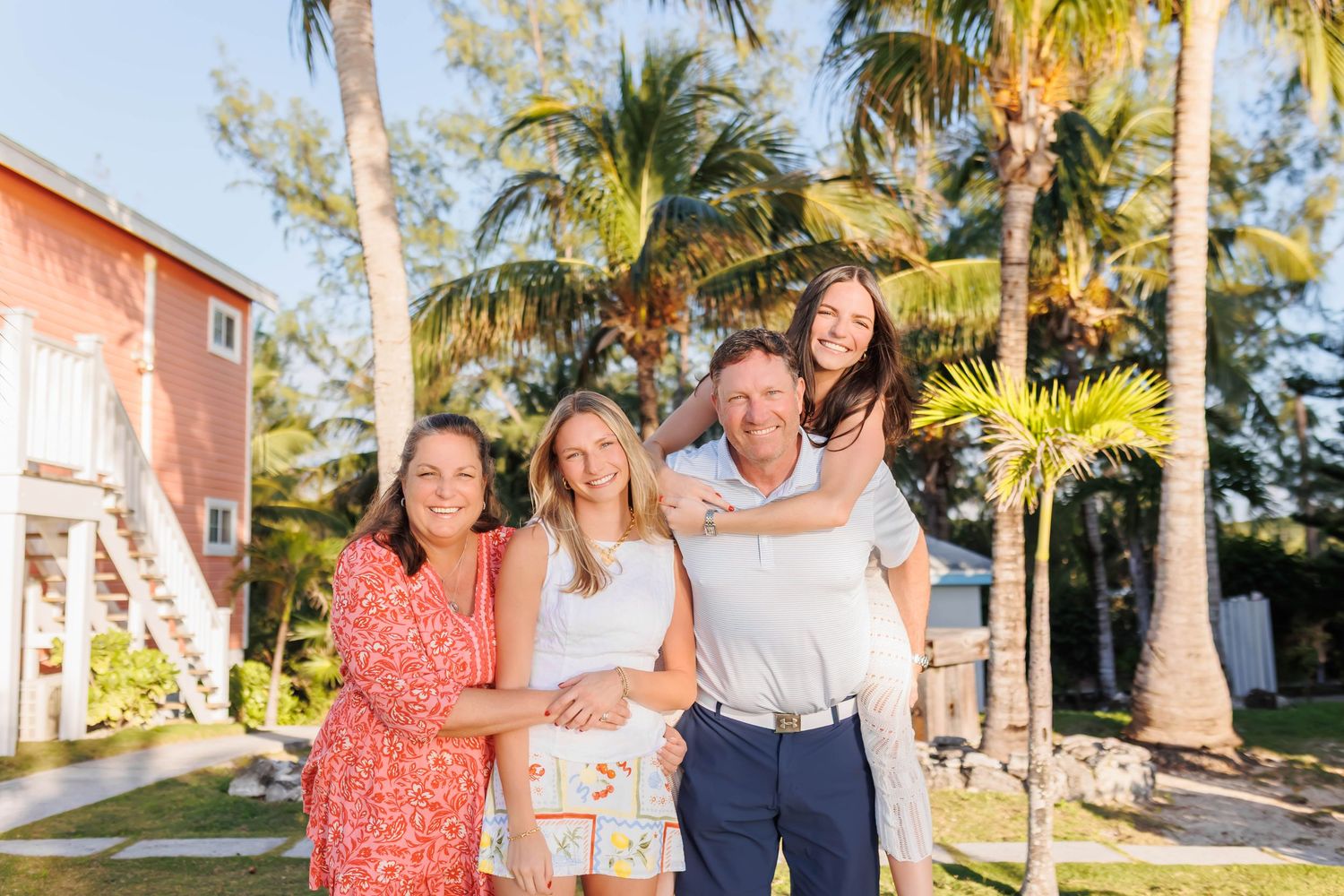 Happy family of four posing together outdoors near palm trees and a pink beach house on a sunny day.