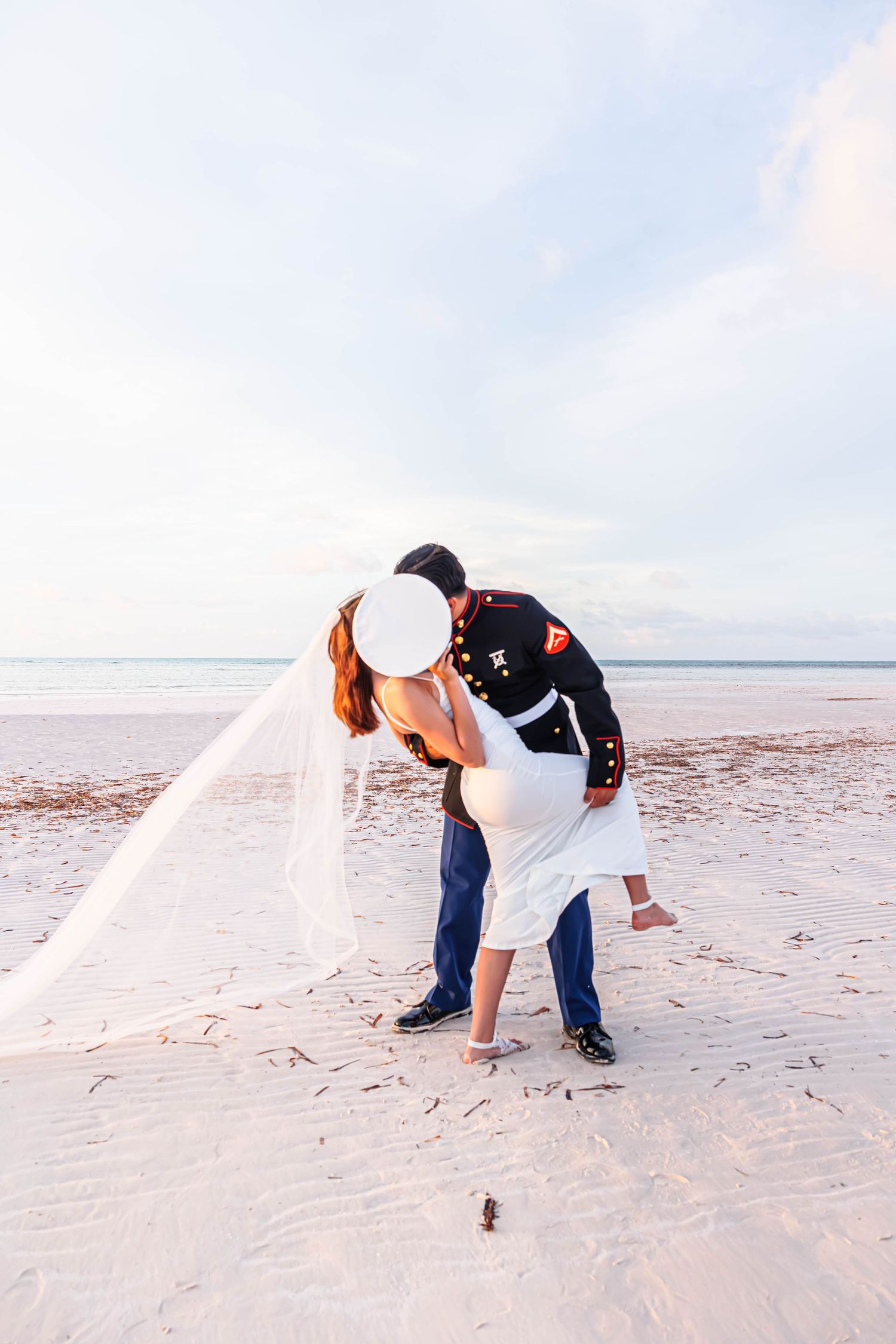 Marine groom dips bride in white dress and veil for romantic beach wedding kiss at sunset.
