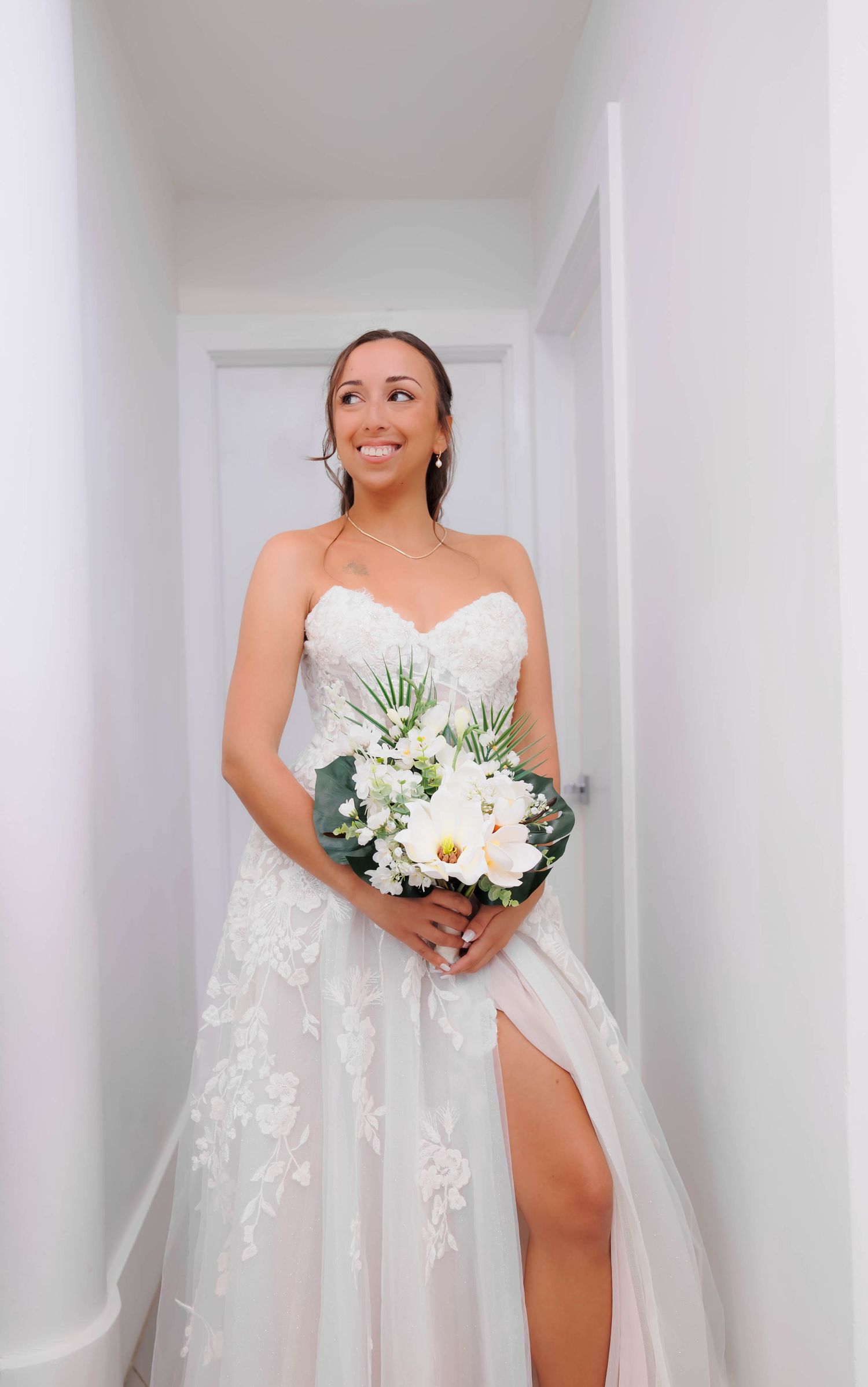 Smiling bride in strapless lace wedding dress with thigh slit holding white bouquet in sunlit corridor.