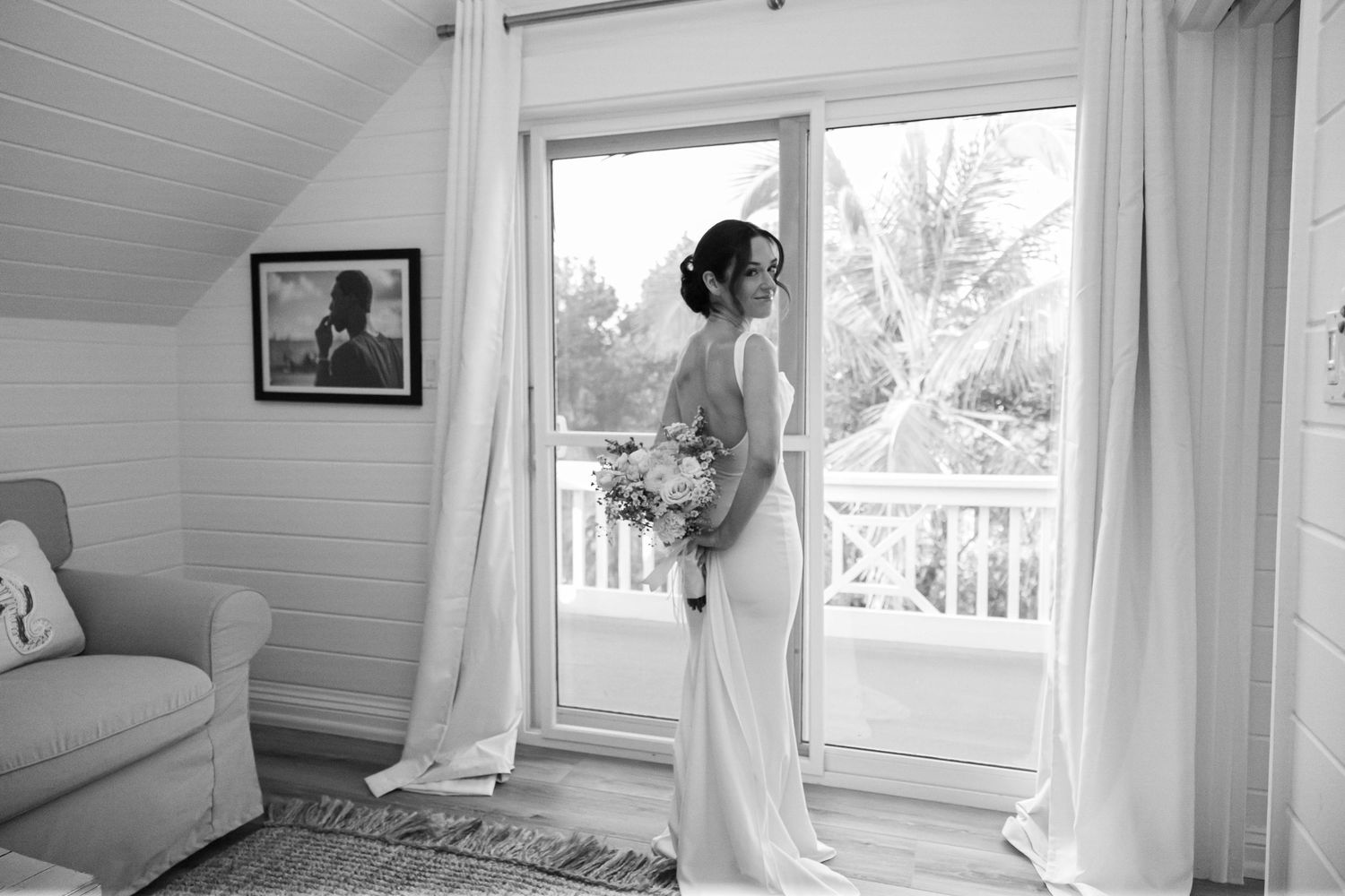 Bride in elegant white gown holding bouquet stands by balcony doors in black and white wedding portrait.