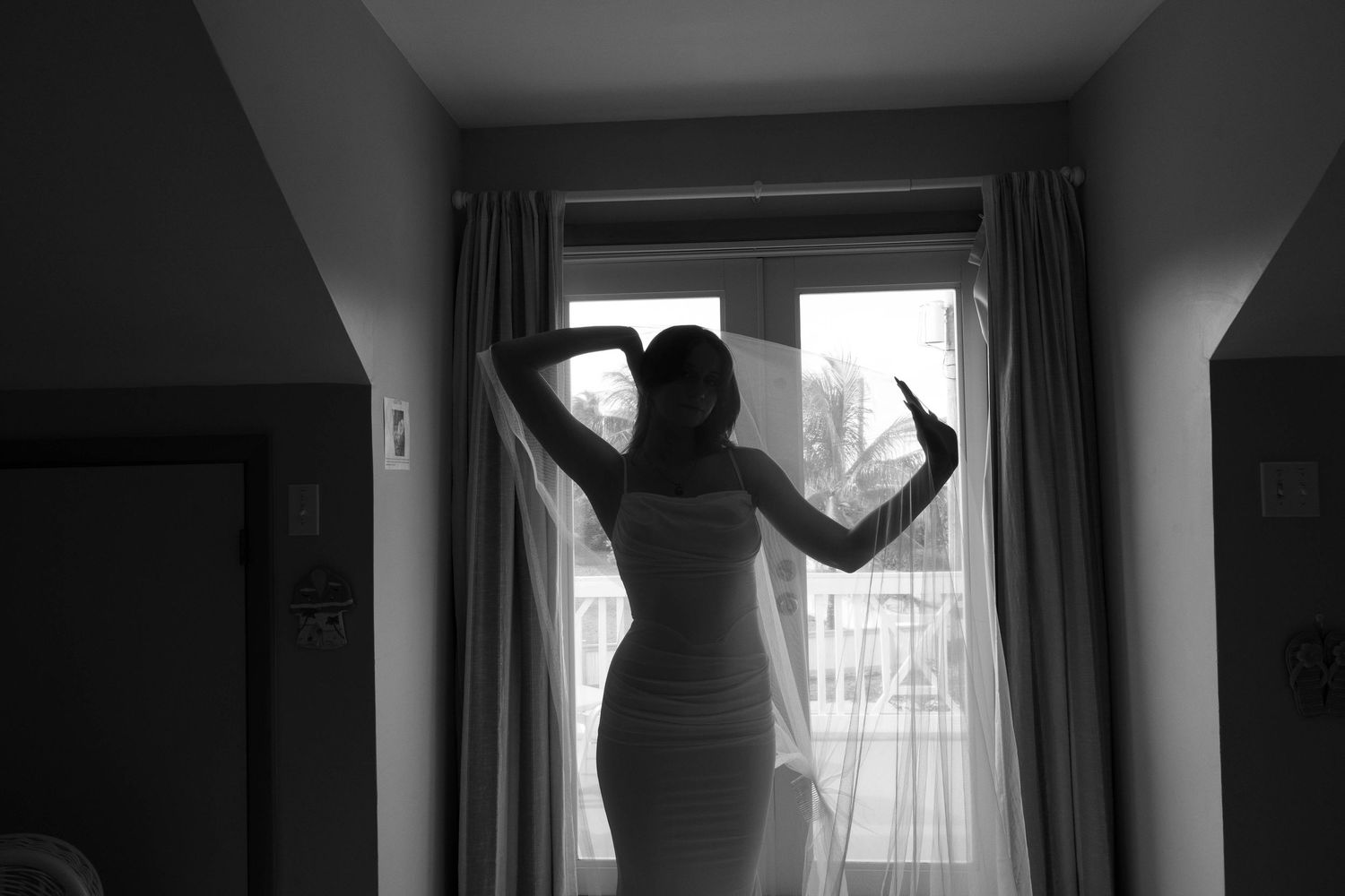 Black and white photo of a woman in a dress standing by a curtained window with arms raised.