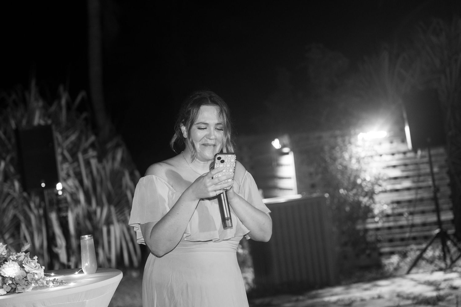 Woman in white dress smiling while holding microphone and phone during outdoor wedding reception speech at night.