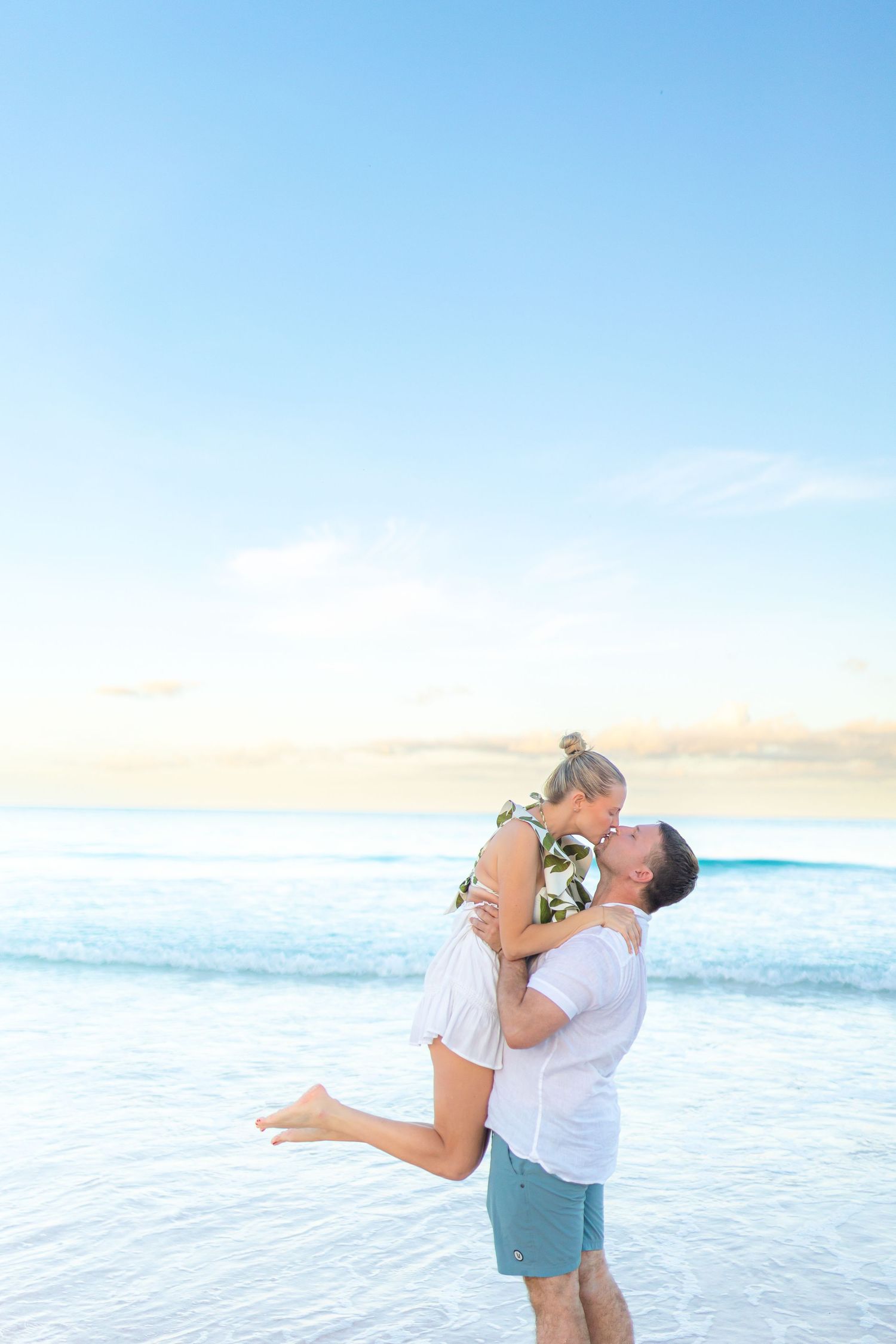 A couple shares a romantic kiss on a tropical beach at sunset, as the man lifts the woman in his arms.