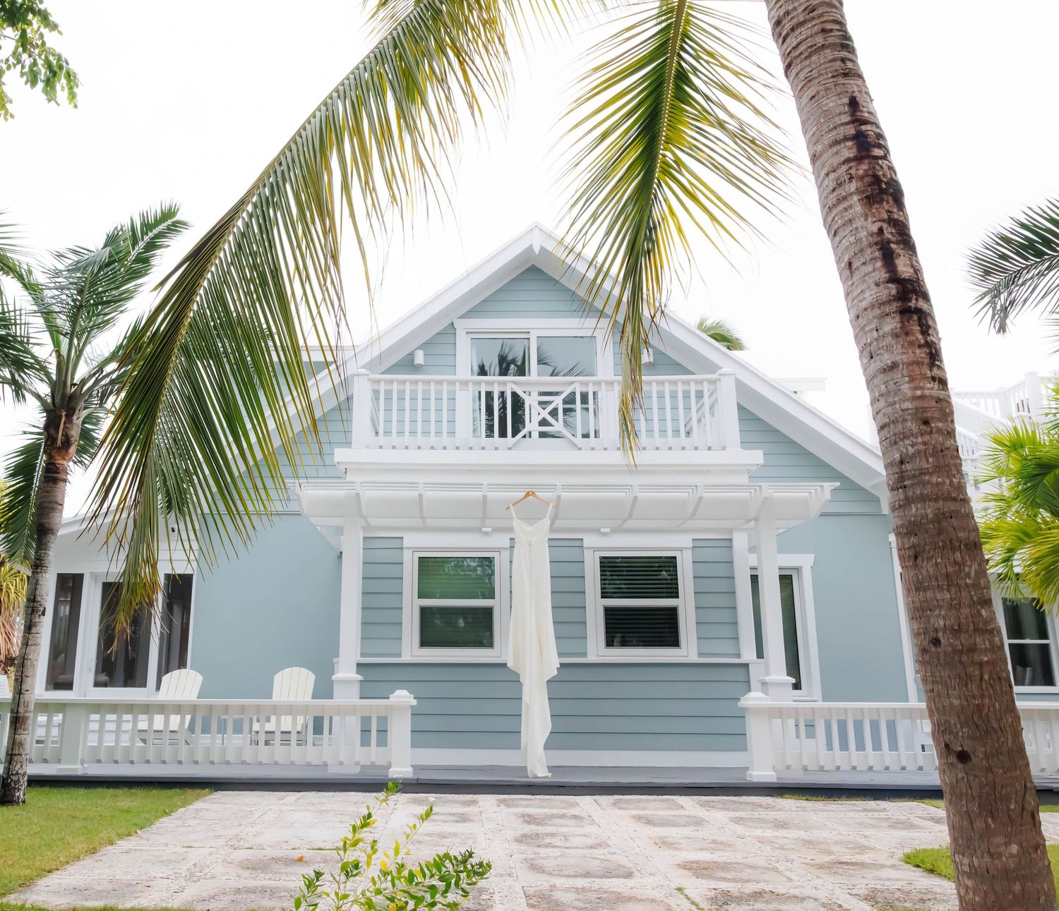 Light blue coastal cottage with white trim, balcony, and wraparound porch surrounded by tropical palm trees.