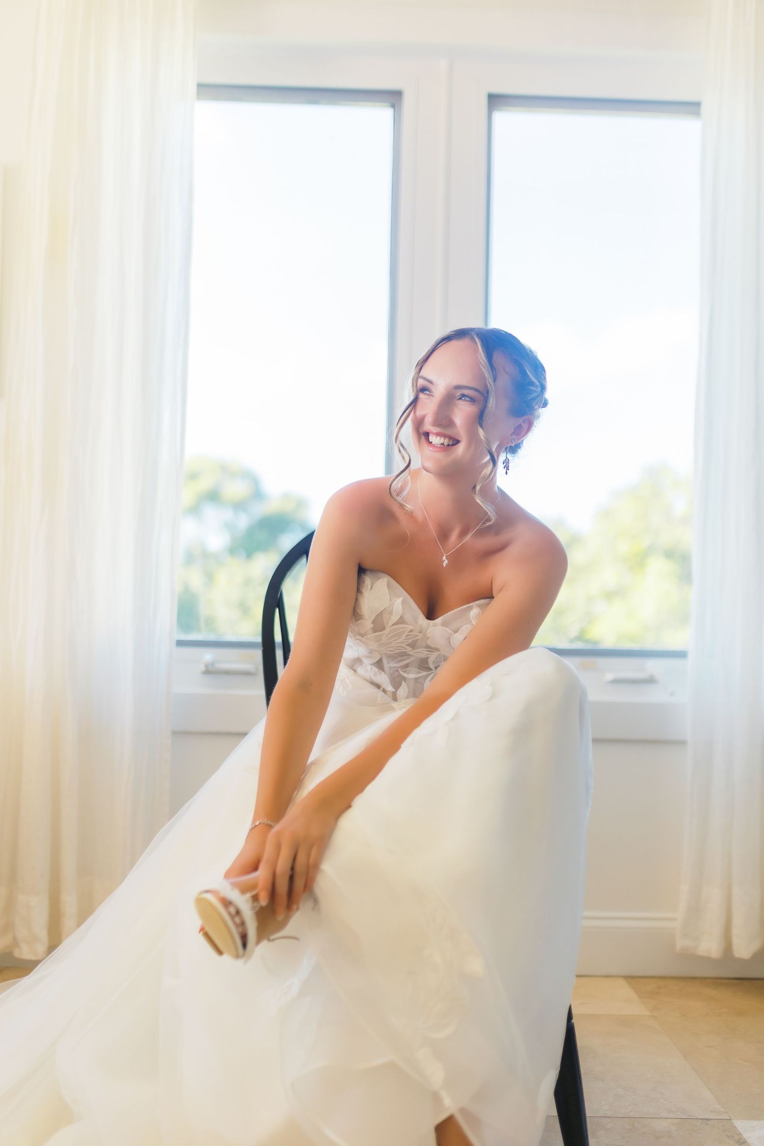 Smiling bride in a strapless lace wedding gown putting on shoes by a bright window.