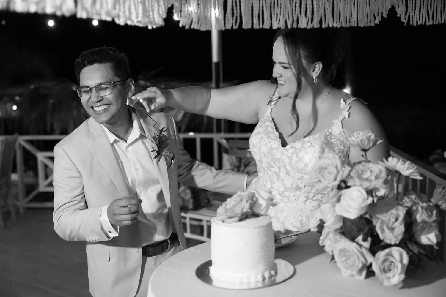 Bride and groom laugh together while cutting their white wedding cake at an outdoor reception.