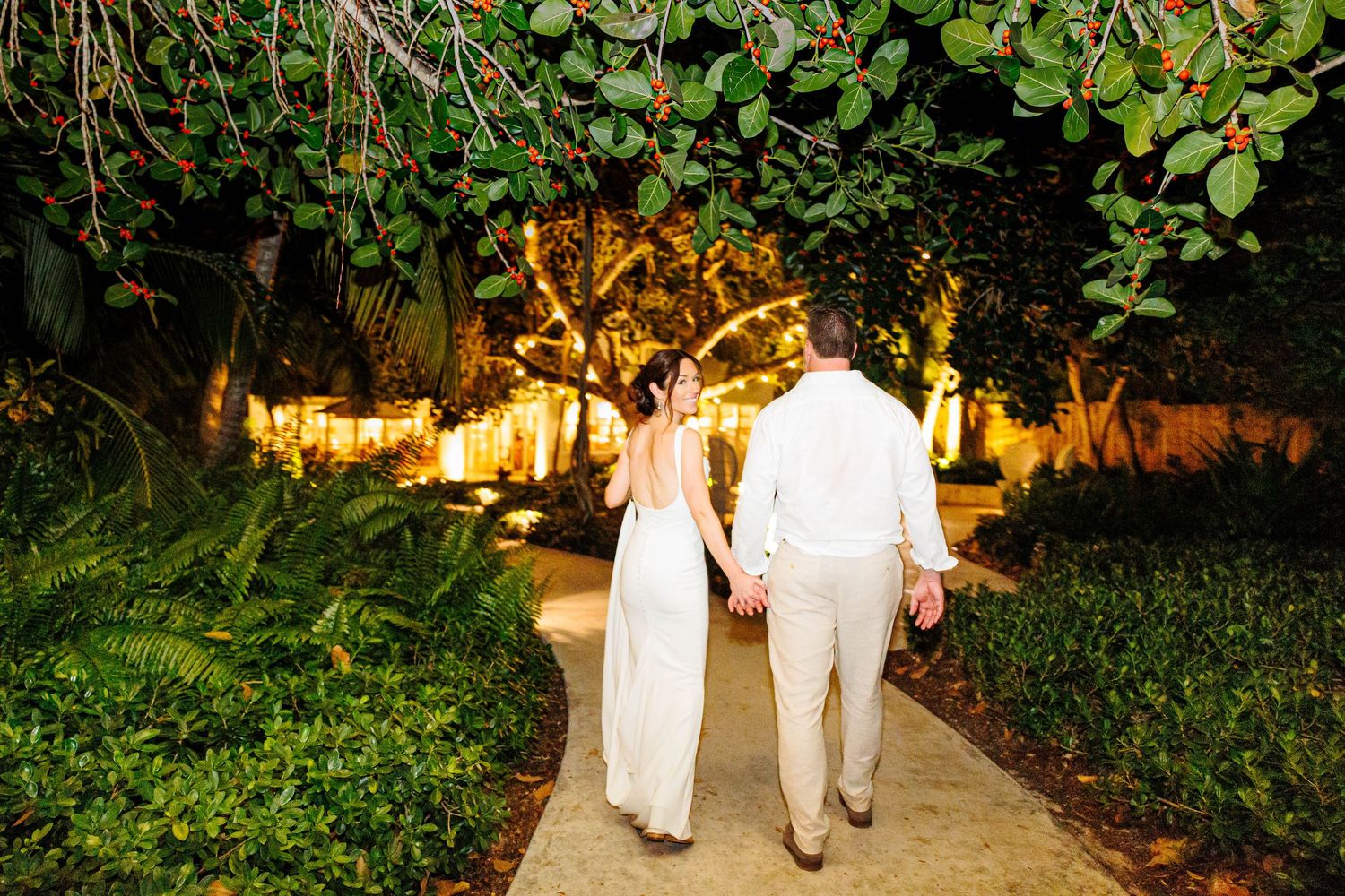 Bride and groom walking hand in hand down a tropical garden path at night, illuminated by warm golden lights.