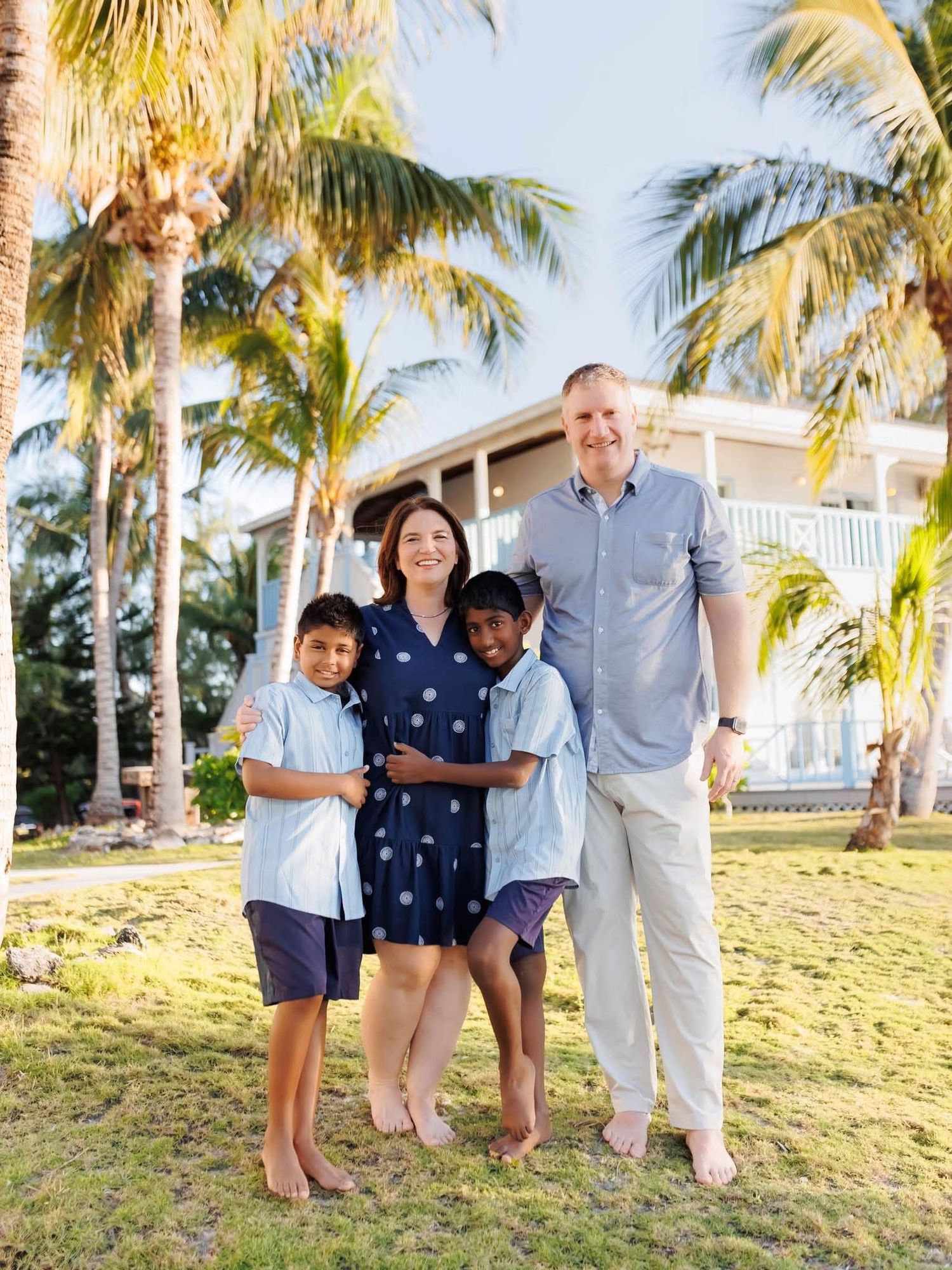 Family of four posing outdoors near palm trees and a tropical house on a sunny day.