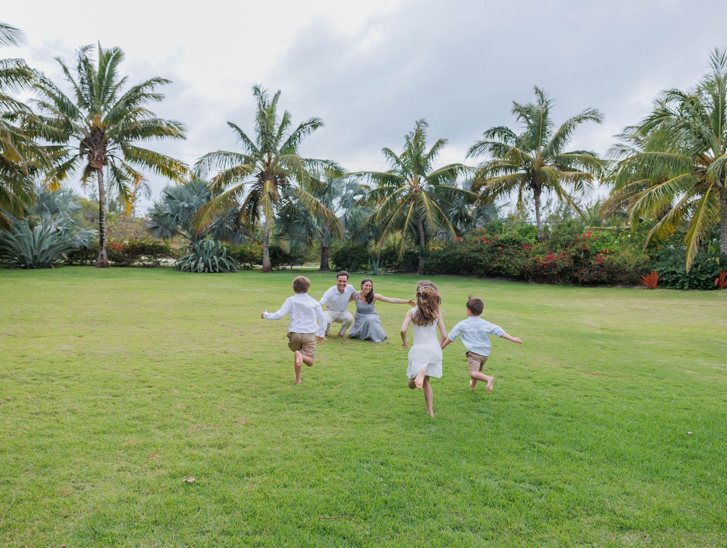 Children in white clothes running toward a woman on a lush green lawn surrounded by tropical palm trees.