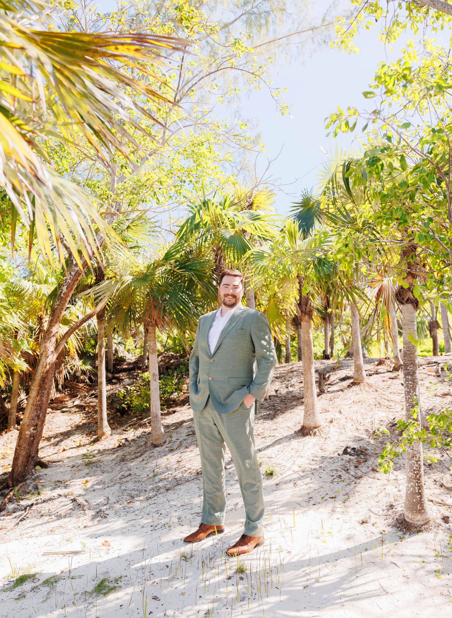 Groom in sage green suit standing on sandy beach path surrounded by tropical palm trees on wedding day.
