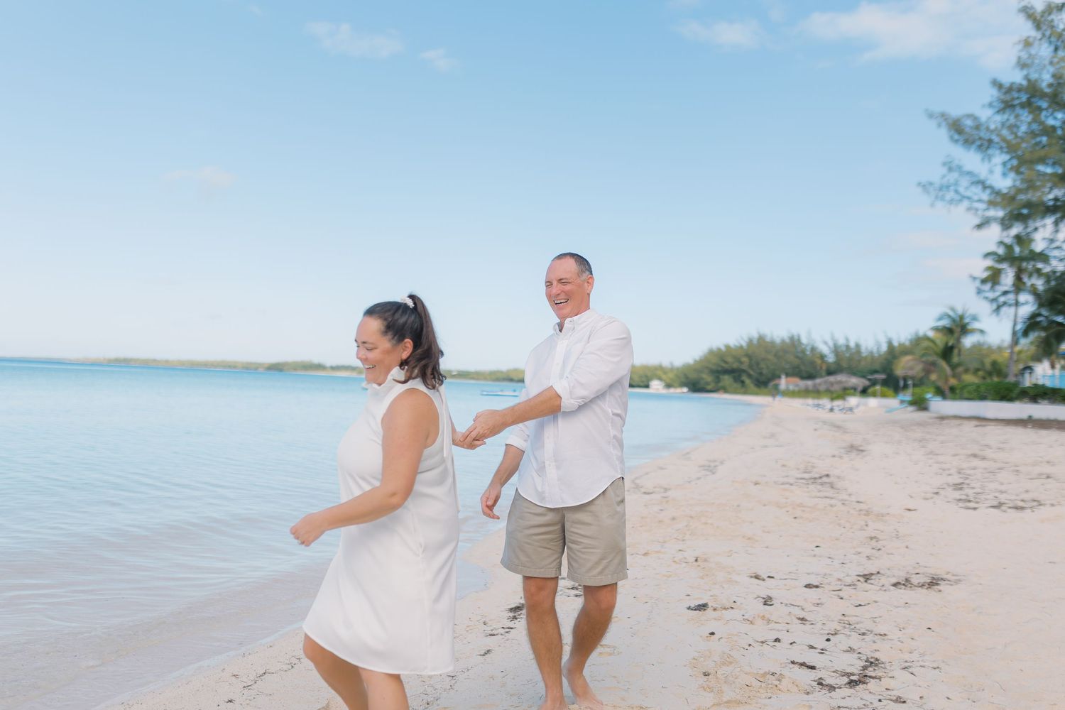 Couple in white outfits holding hands and walking barefoot on a tropical sandy beach with calm turquoise waters.