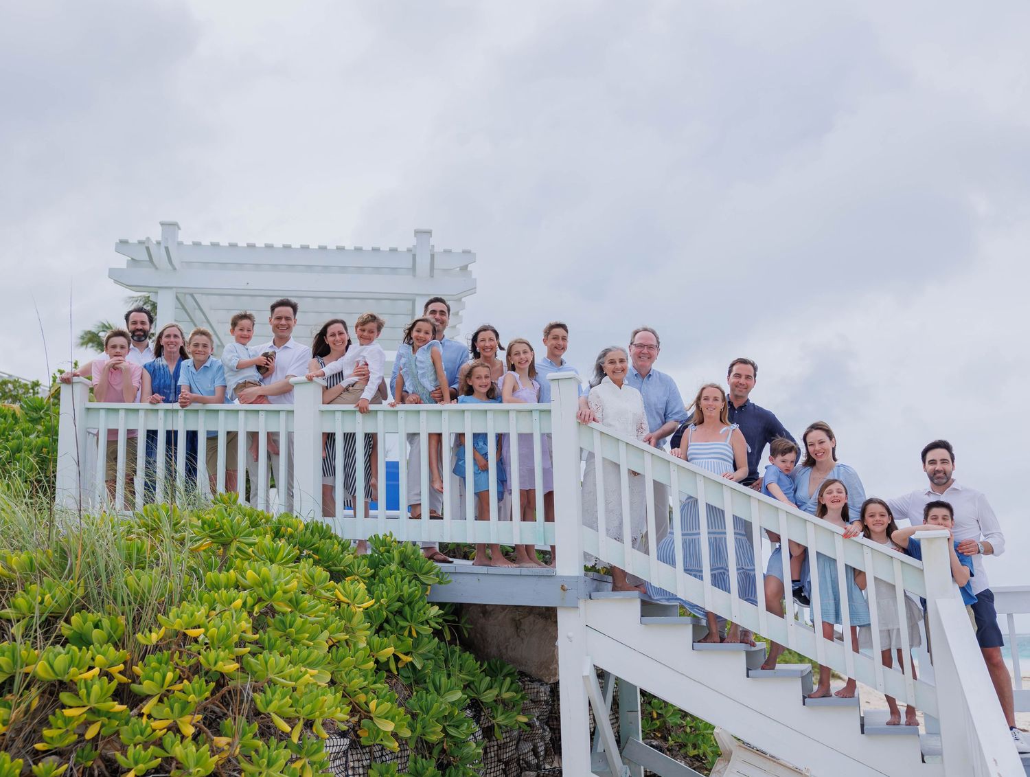 Large family group posing on white beachside staircase deck with lush greenery and cloudy sky backdrop.