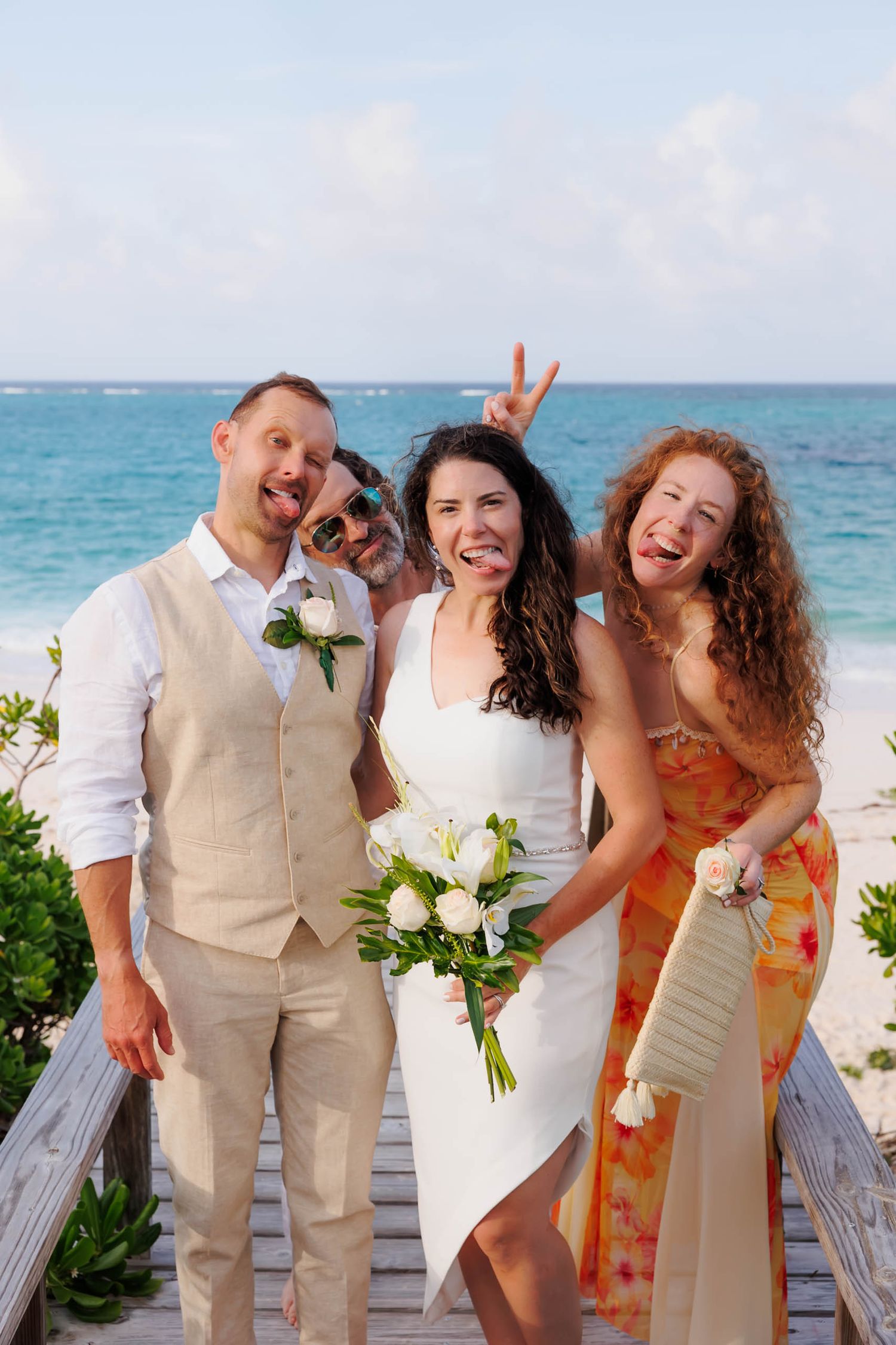 Wedding couple and guests laugh on beach boardwalk, bride holding white bouquet, turquoise ocean backdrop.