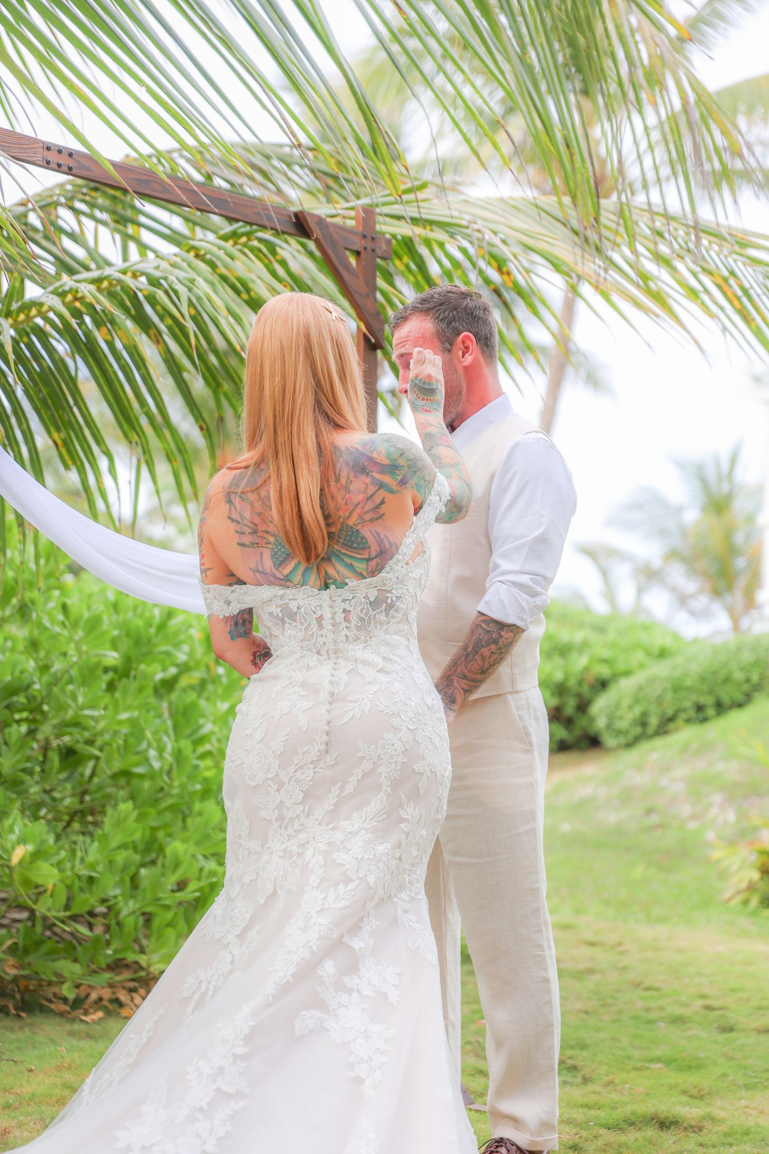 Tattooed bride in lace mermaid gown and groom share intimate moment under palm tree arch.