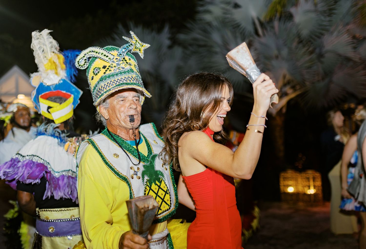wedding day at potlatch Eleuthera  carnival performer in festive costume stands beside a woman in red dress at a tropical night event.