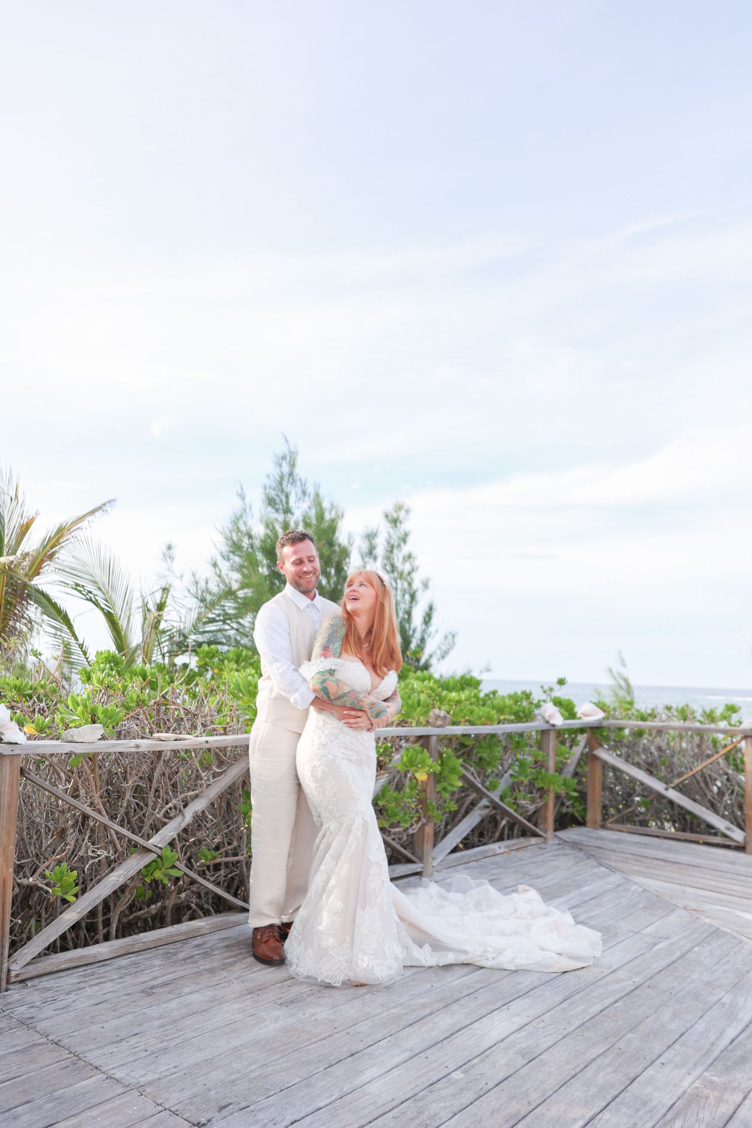 Smiling bride and groom posing on a rustic wooden deck with tropical trees behind them.