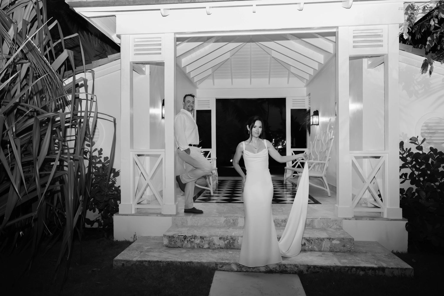 Black and white photo of a bride and groom posing at the entrance of an elegant white wedding venue at night.