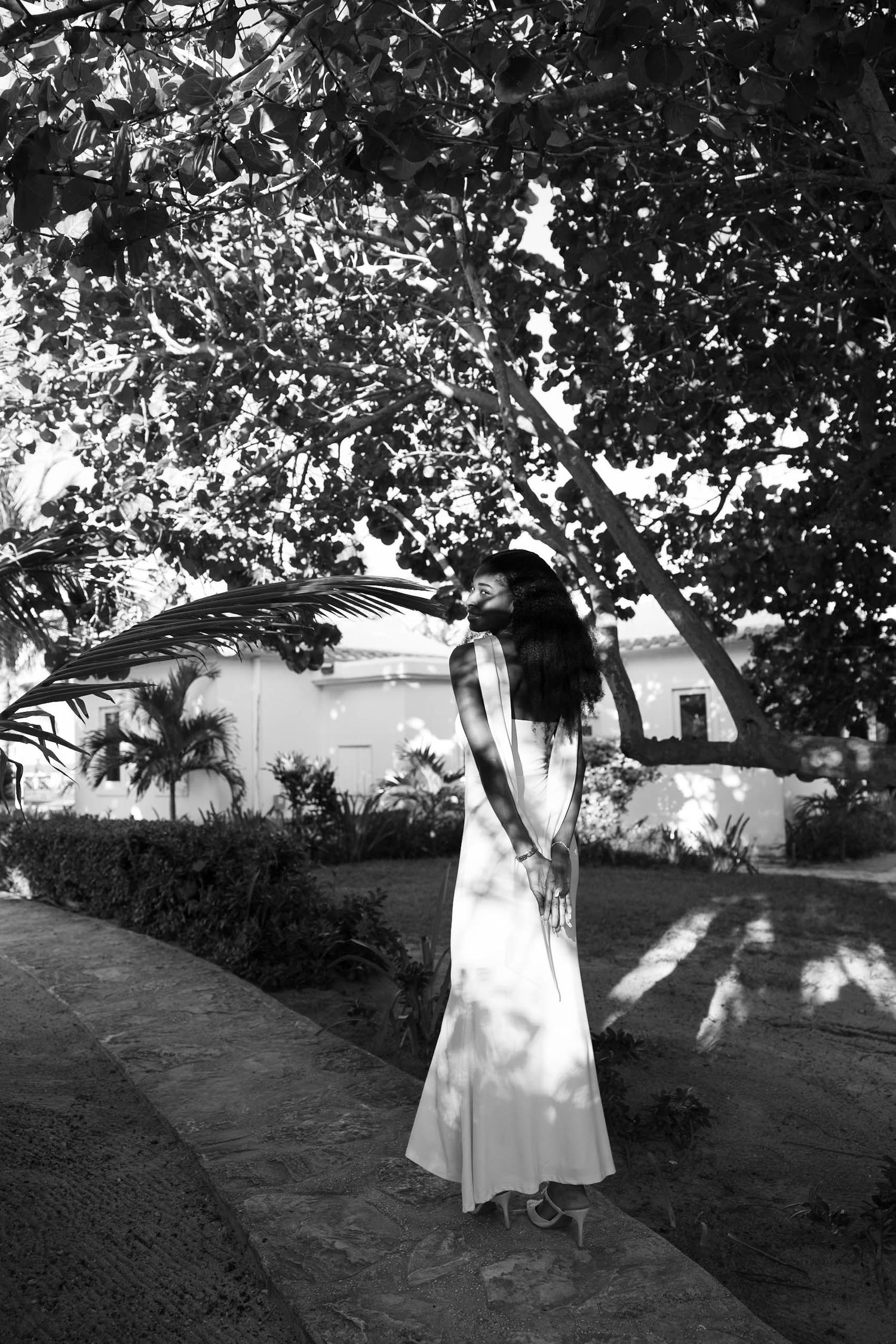 Black and white photo of a woman in a white dress standing under a tree in a garden.