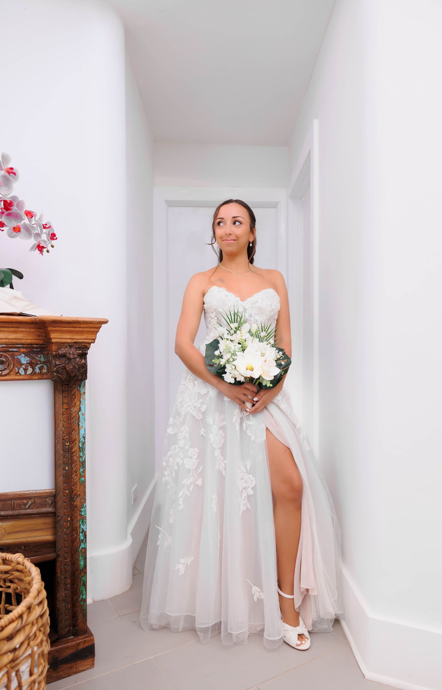 Bride in strapless lace wedding gown with slit holding white floral bouquet standing in bright hallway.