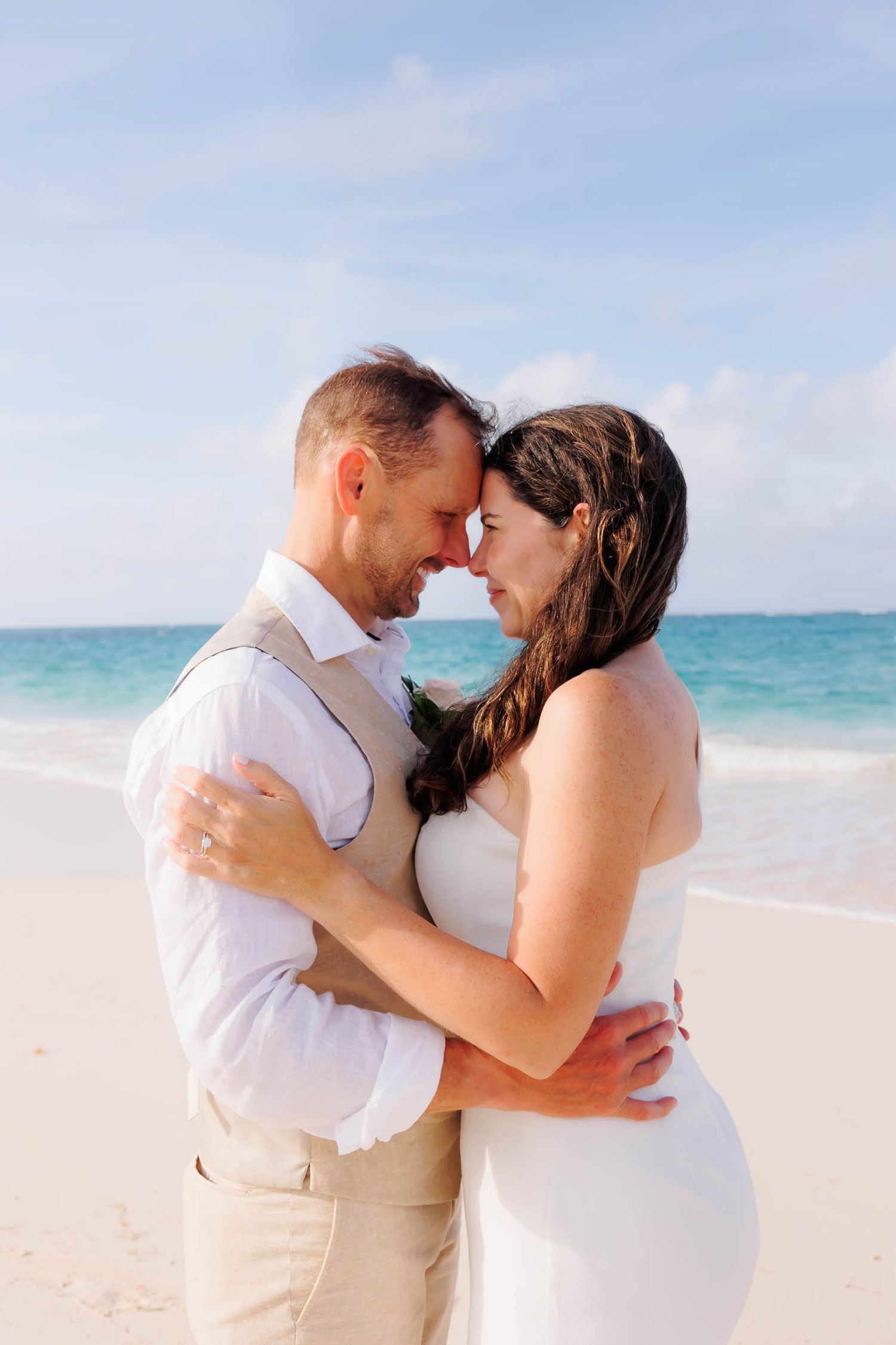 Couple in wedding attire embracing on a tropical beach with turquoise ocean backdrop.