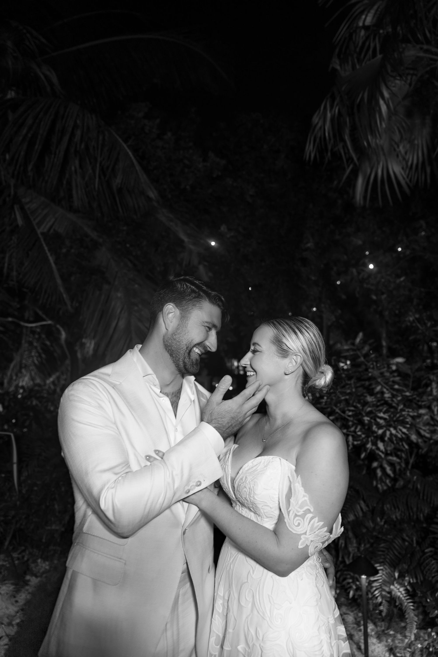 Black and white photo of laughing newlyweds sharing joyful moment together in tropical garden at night.