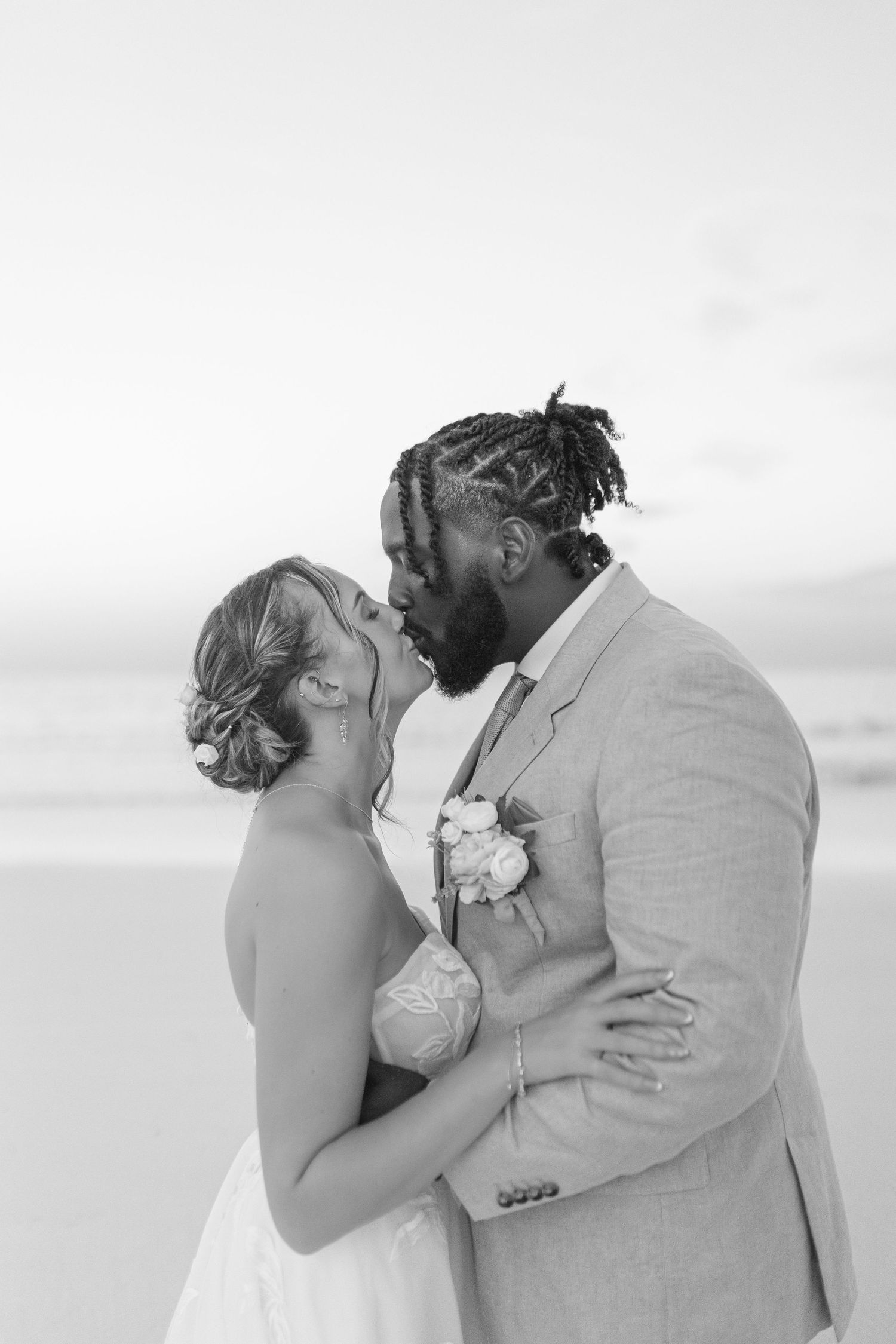 Black and white photo of an interracial couple sharing a kiss on the beach at sunset.