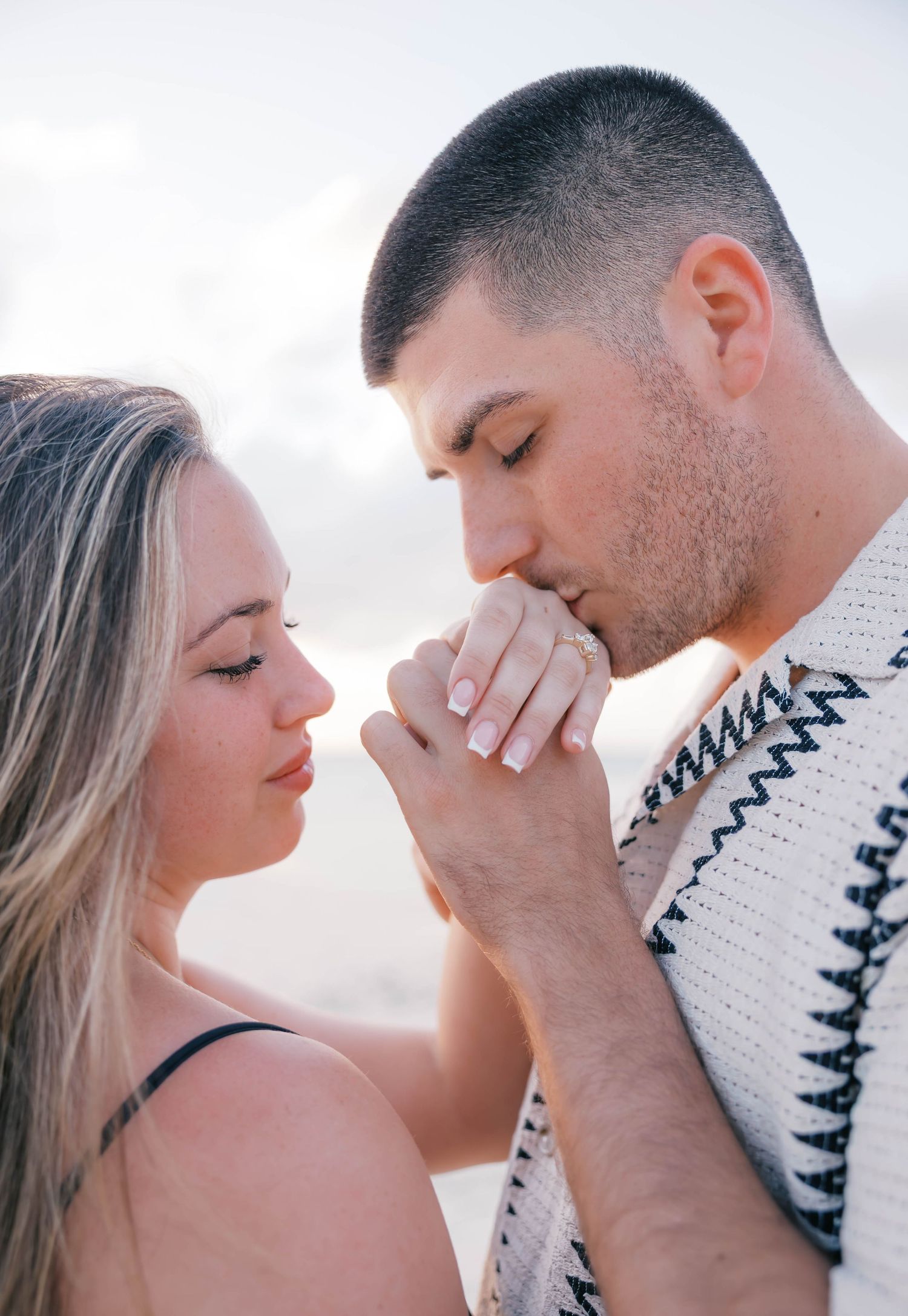 A man tenderly kisses a woman's hand displaying an engagement ring on a romantic beach setting.