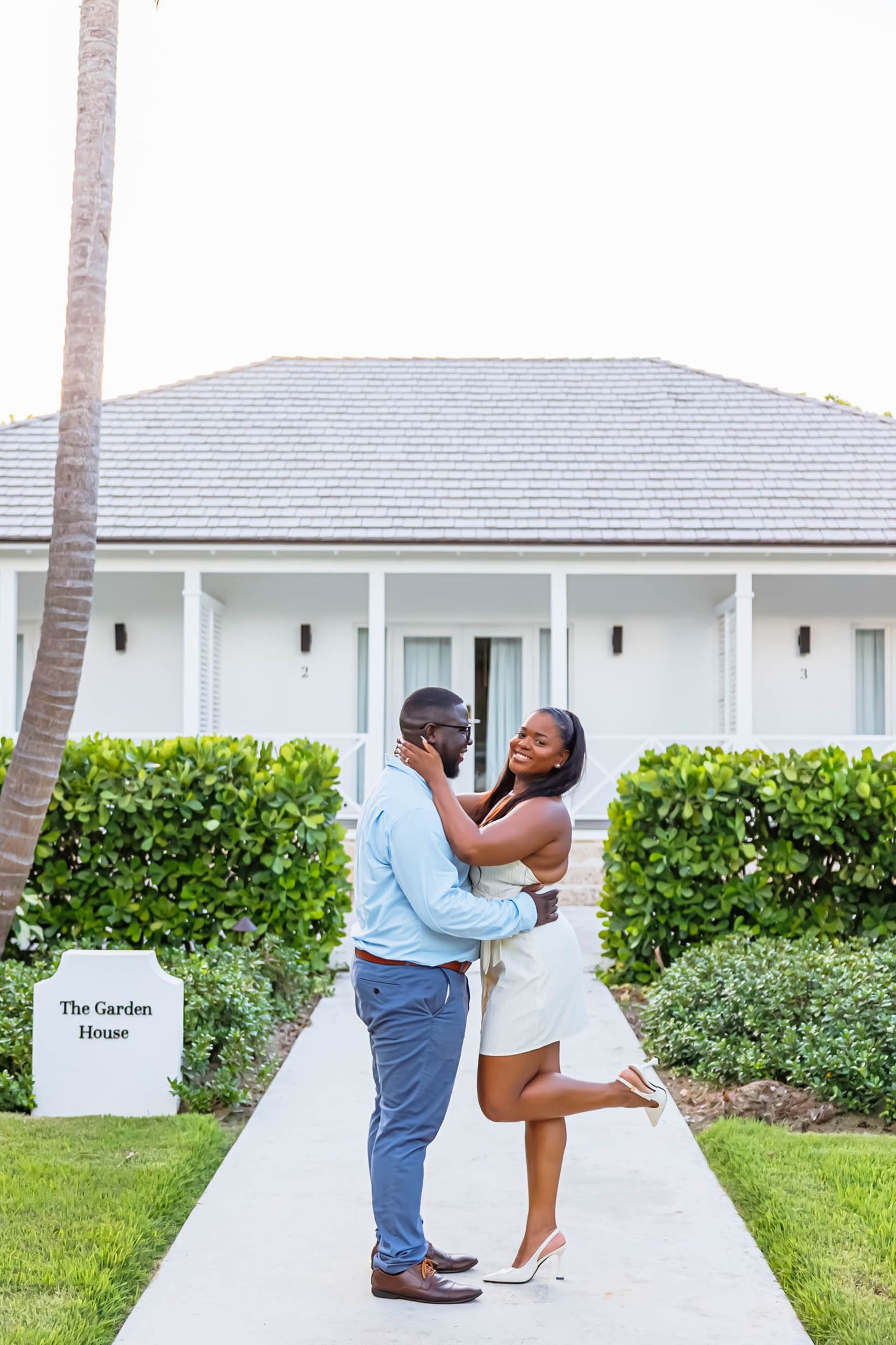 A couple embraces and smiles on a garden path outside The Garden House, dressed in white and blue attire.