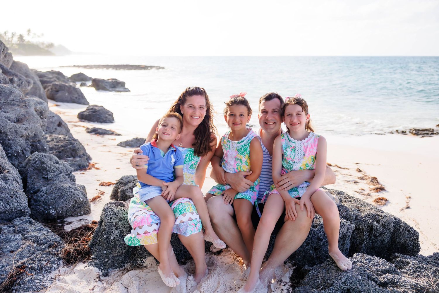 A family of five poses on rocky shoreline in colorful matching outfits with ocean waves and sandy beach in background.