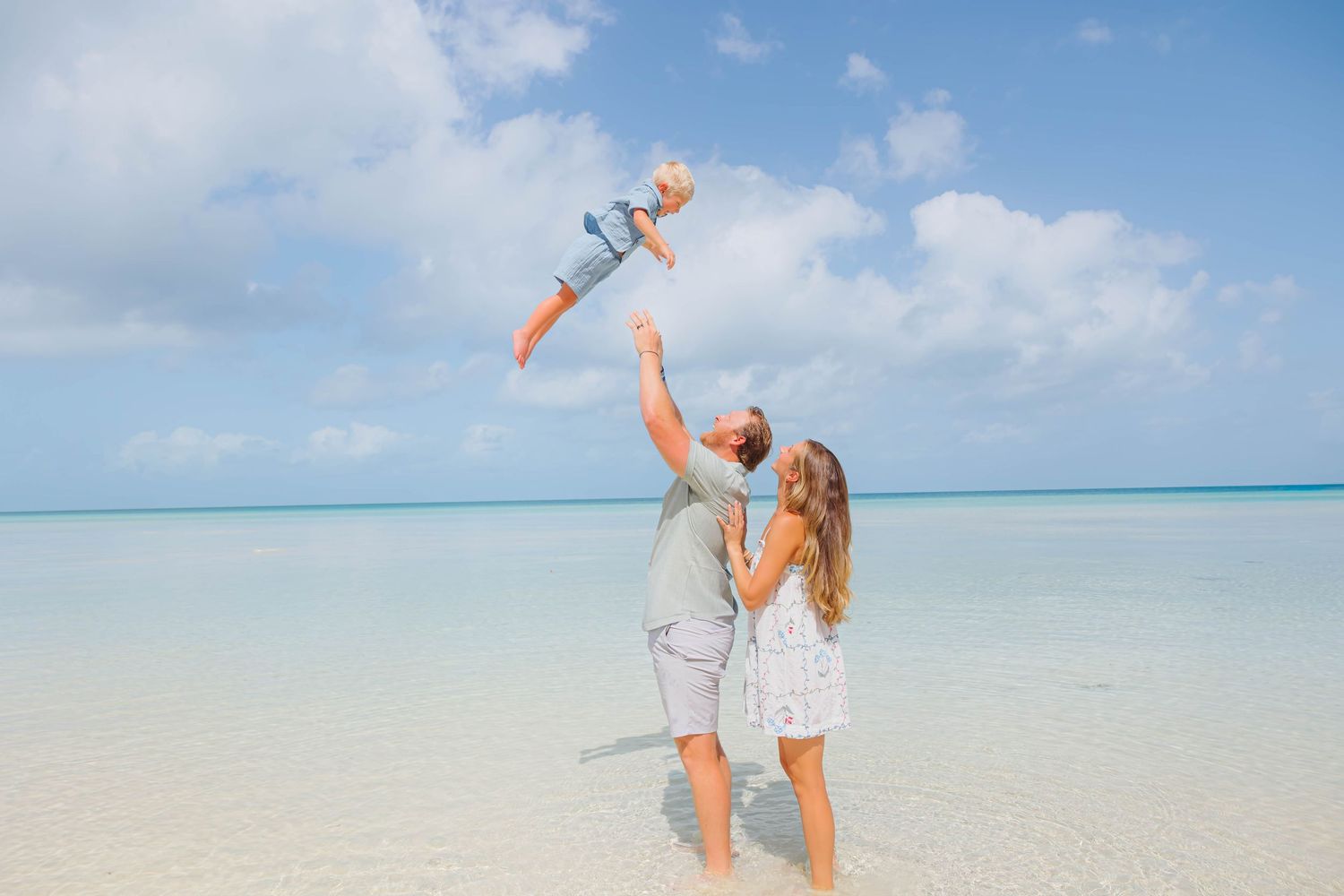 Happy family on tropical beach, parents lifting baby in the air with clear turquoise water and blue sky.