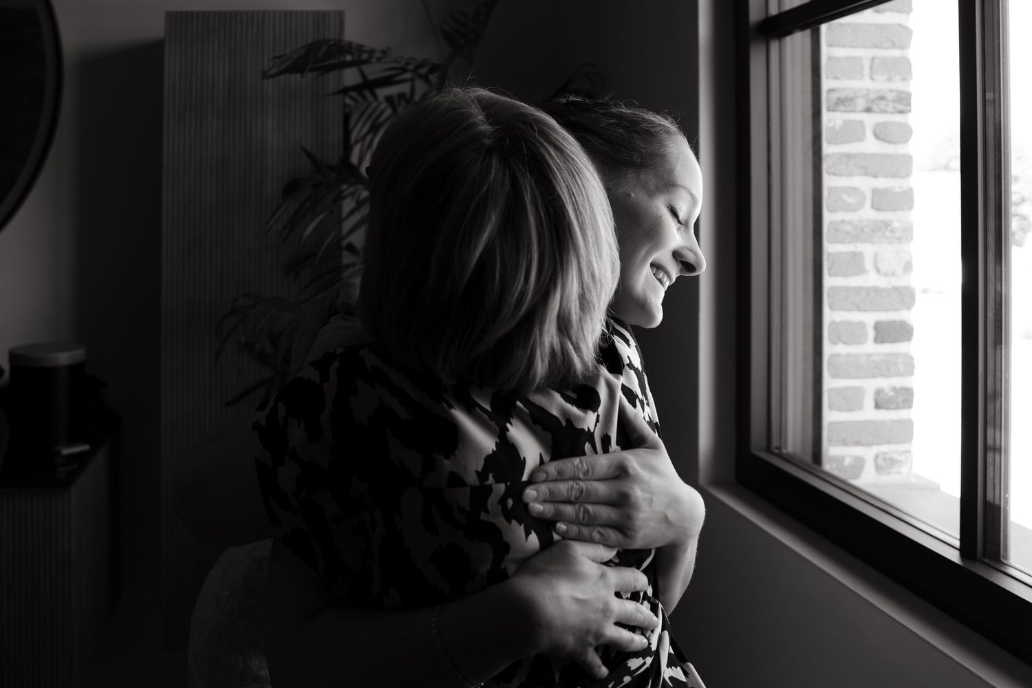 Black and white photo of two people sharing a warm embrace near a window with soft natural light.