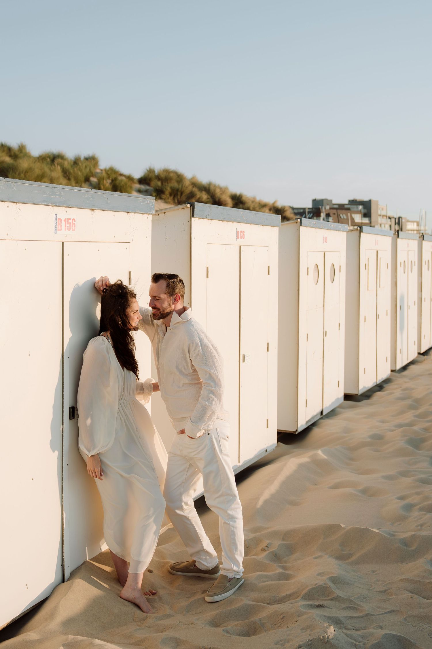 Romantic couple in white outfits leaning against beach cabins on sandy shore at golden hour.