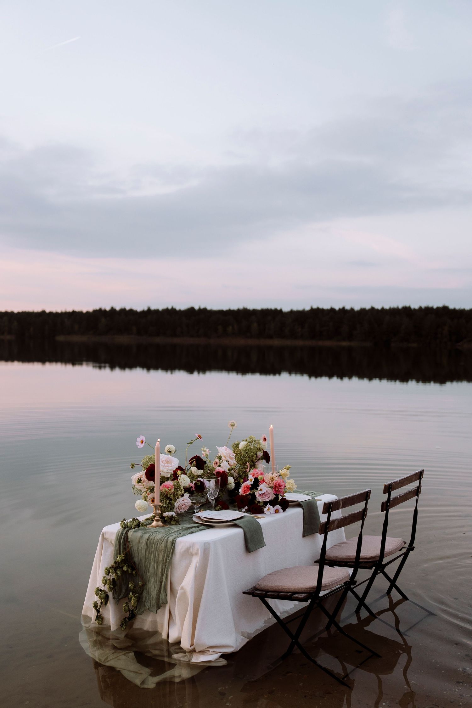 Romantic outdoor dining table set in a calm lake at dusk, adorned with floral arrangements and candles.