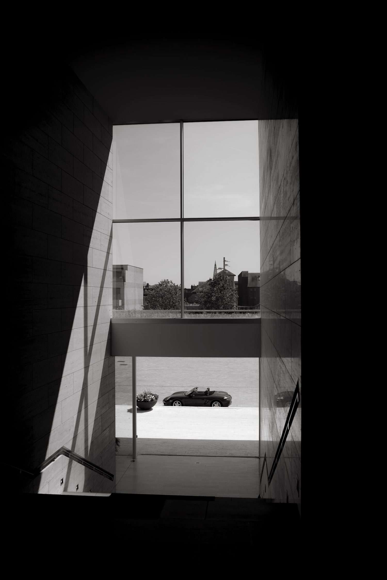 Black and white architectural interior view through large glass windows showing a convertible car parked outside a modern building.
