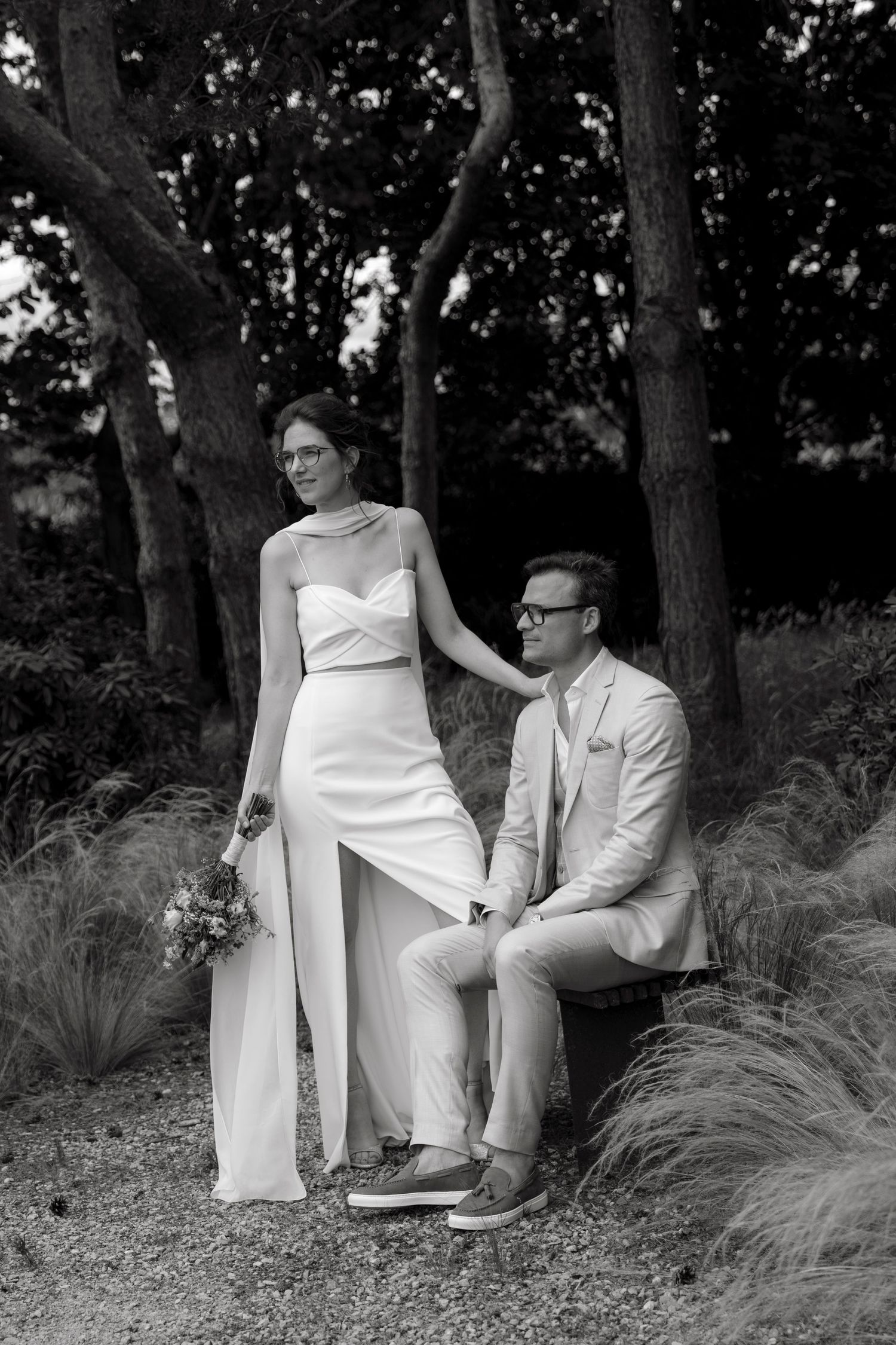 Black and white wedding photo of bride in white pantsuit holding bouquet with groom in light suit seated outdoors.