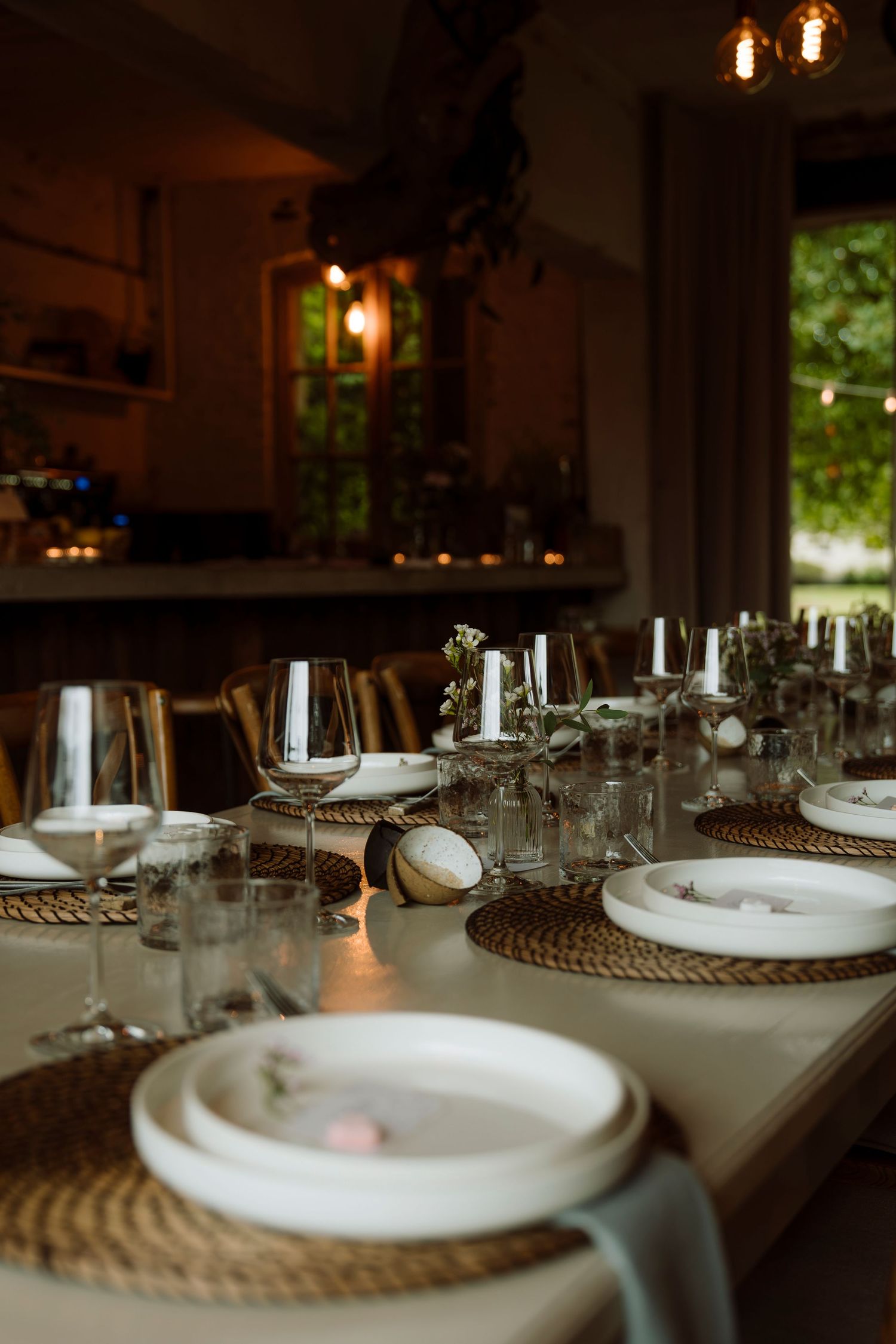 Elegant dinner table setting with wine glasses, white plates, woven placemats, and candles in a warmly lit dining room.