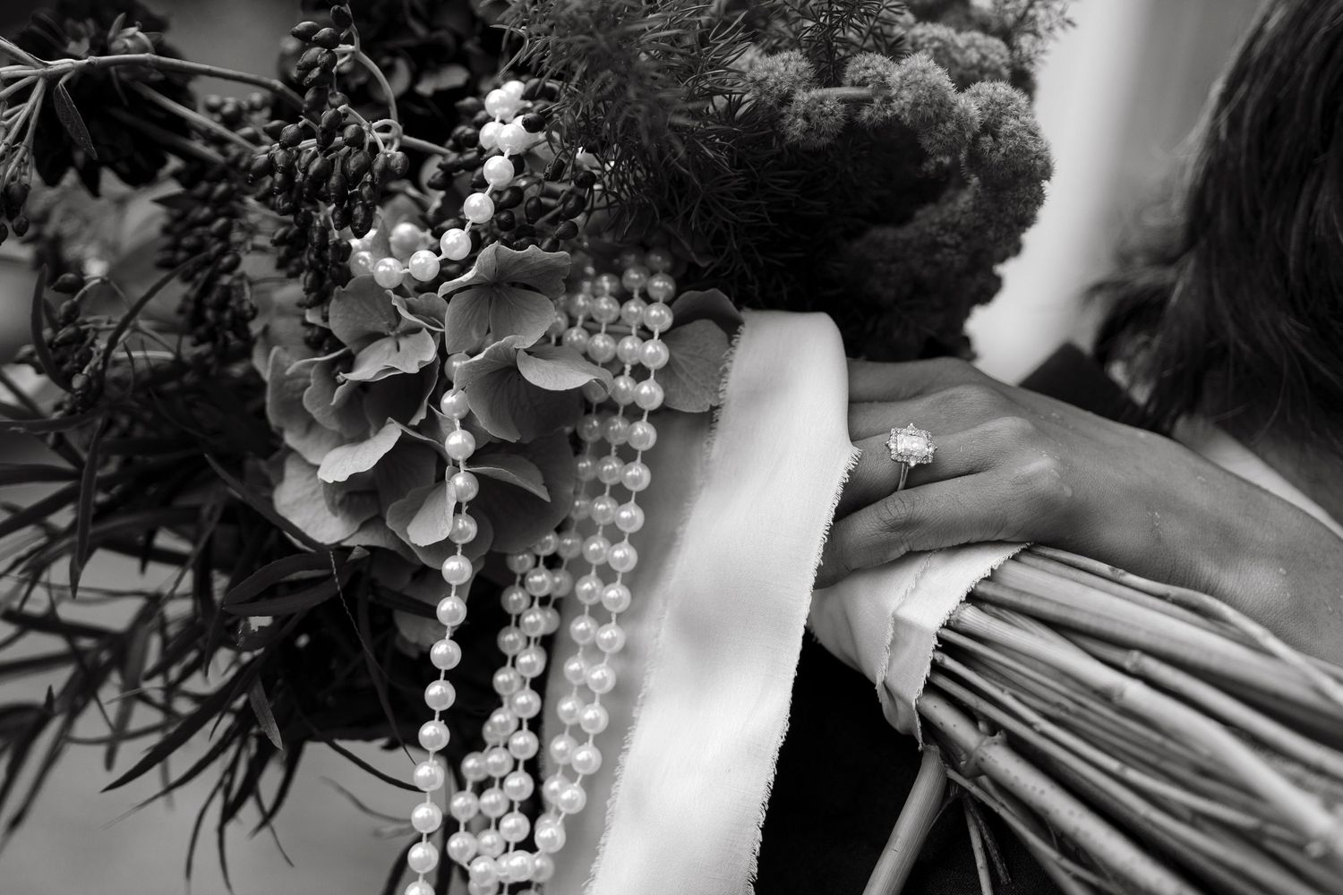 Black and white photo of bride holding floral bouquet with pearl necklace and diamond engagement ring.