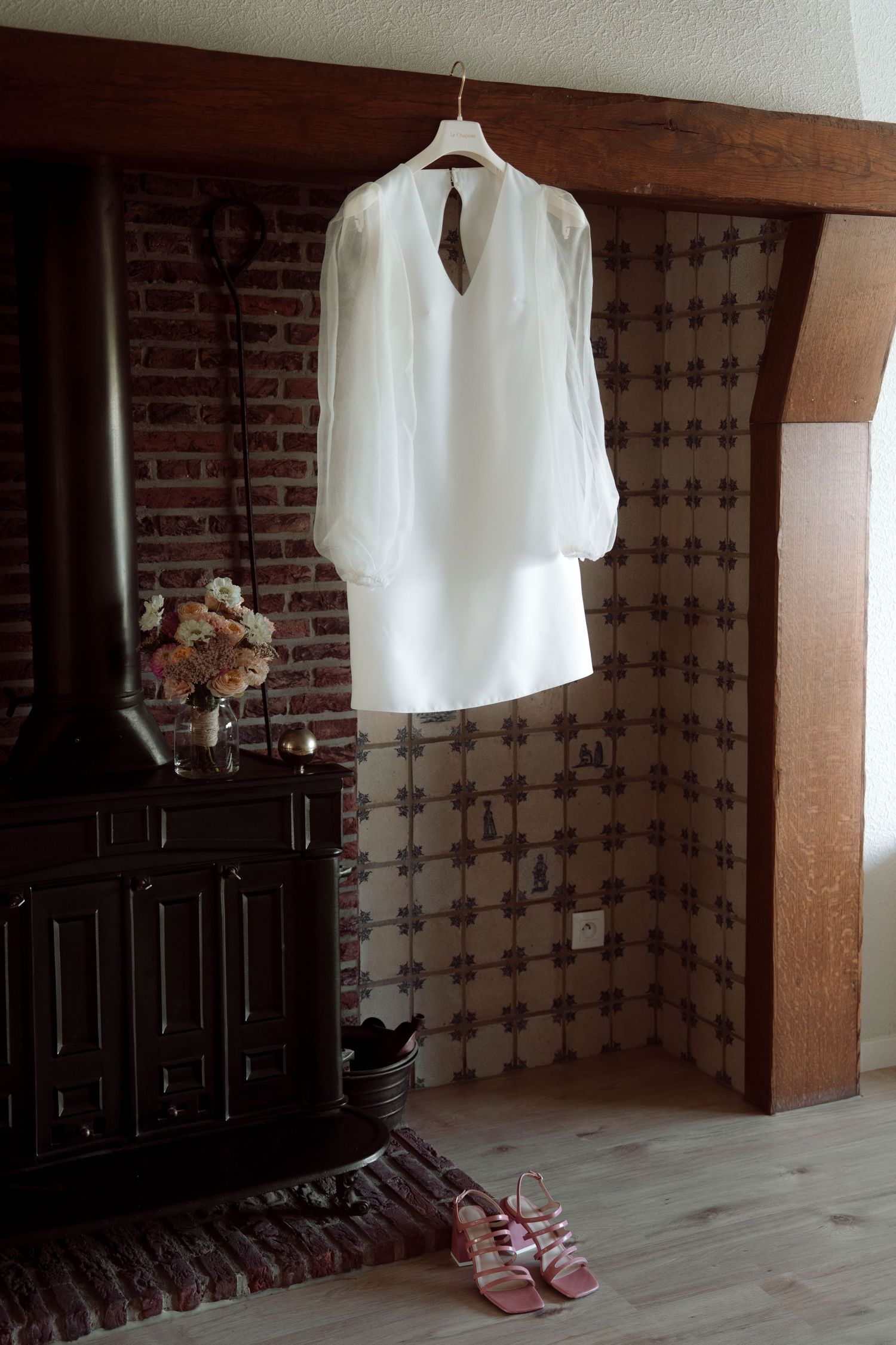 Elegant white long-sleeve wedding dress hanging on hanger near rustic brick fireplace with pink heels below.